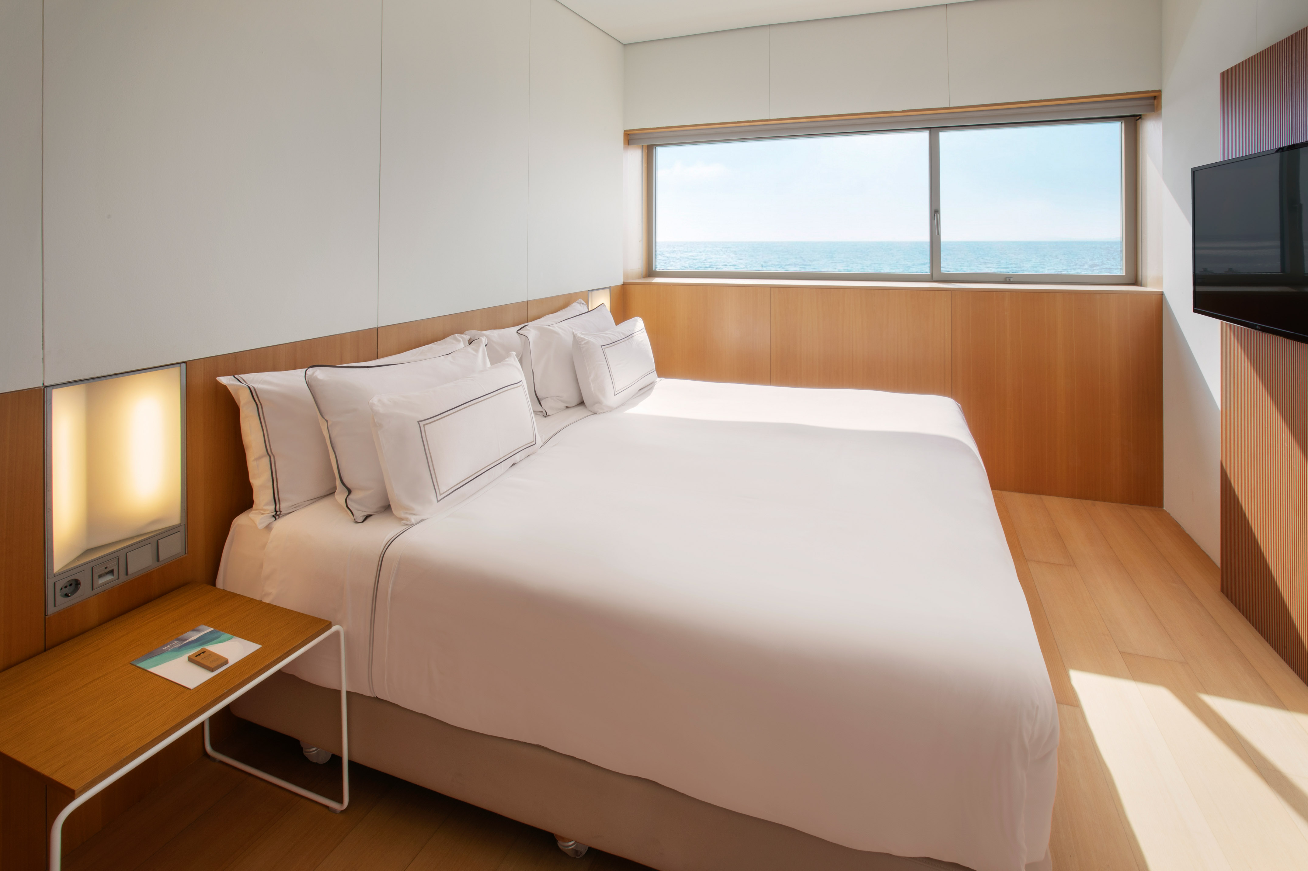 Modern hotel bedroom with ocean view, white bed, and wood accents.