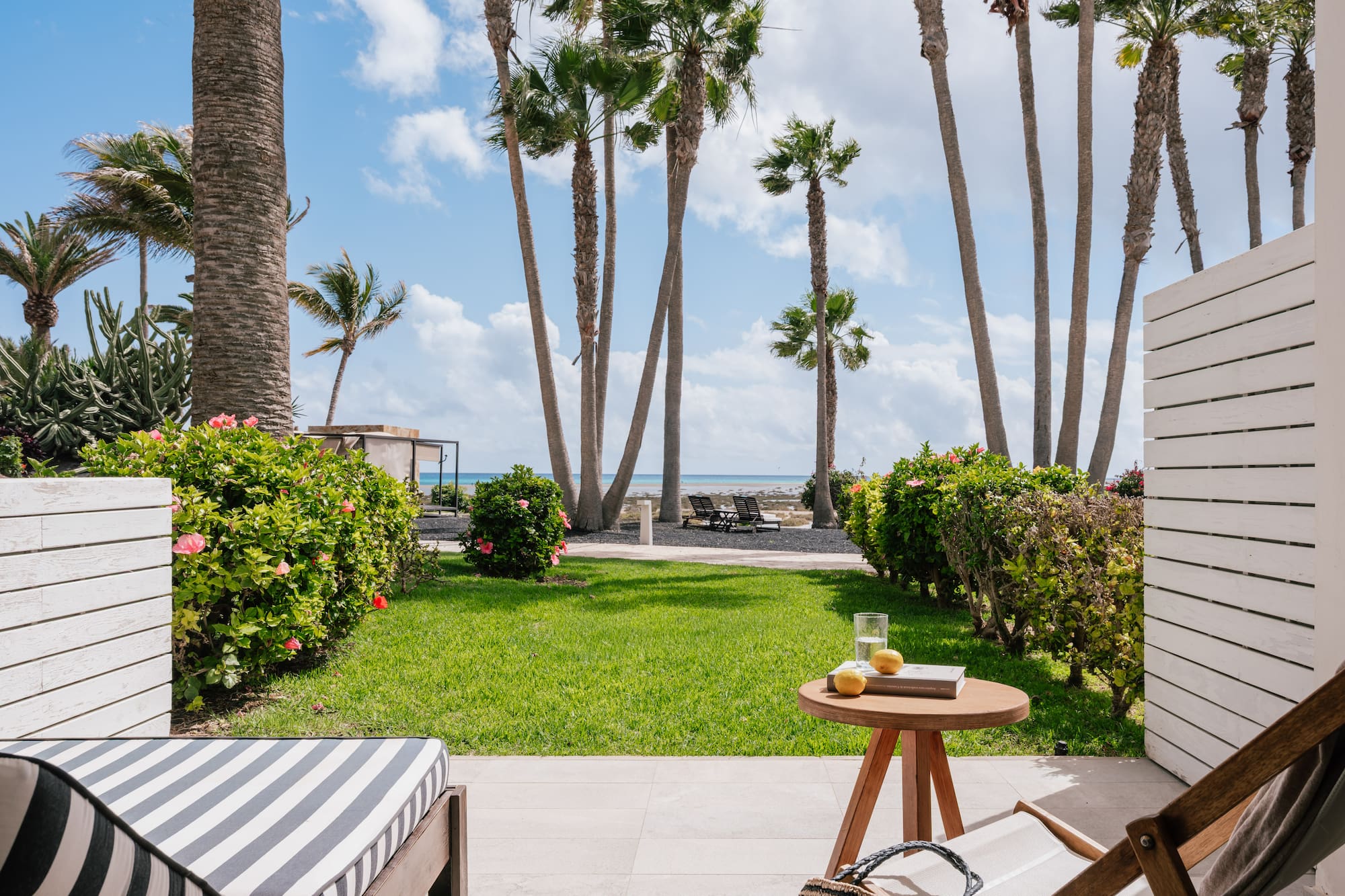 Terraza de resort con palmeras, jardín, tumbonas y mar azul.