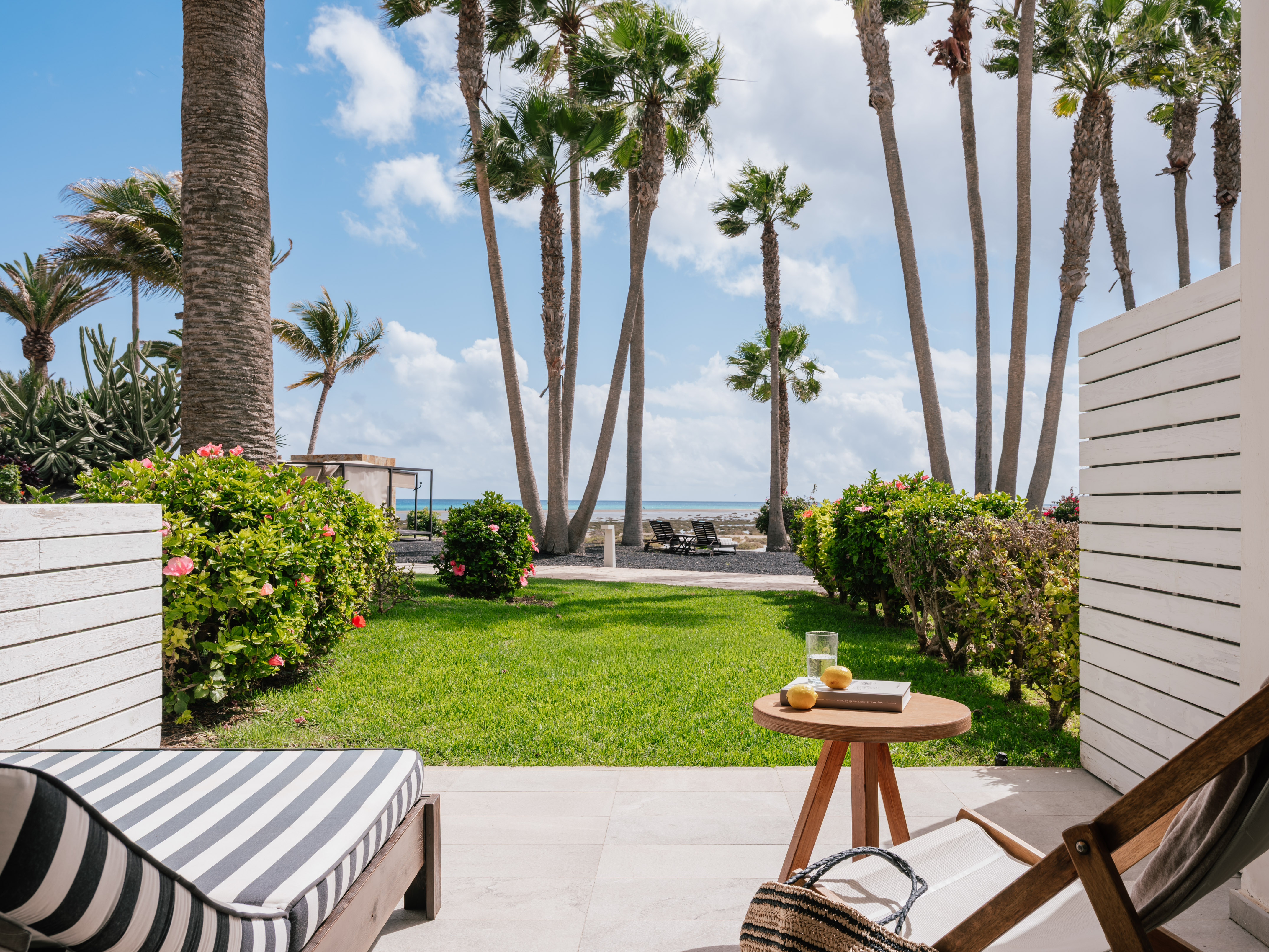Terraza de resort con palmeras, jardín, tumbonas y mar azul.
