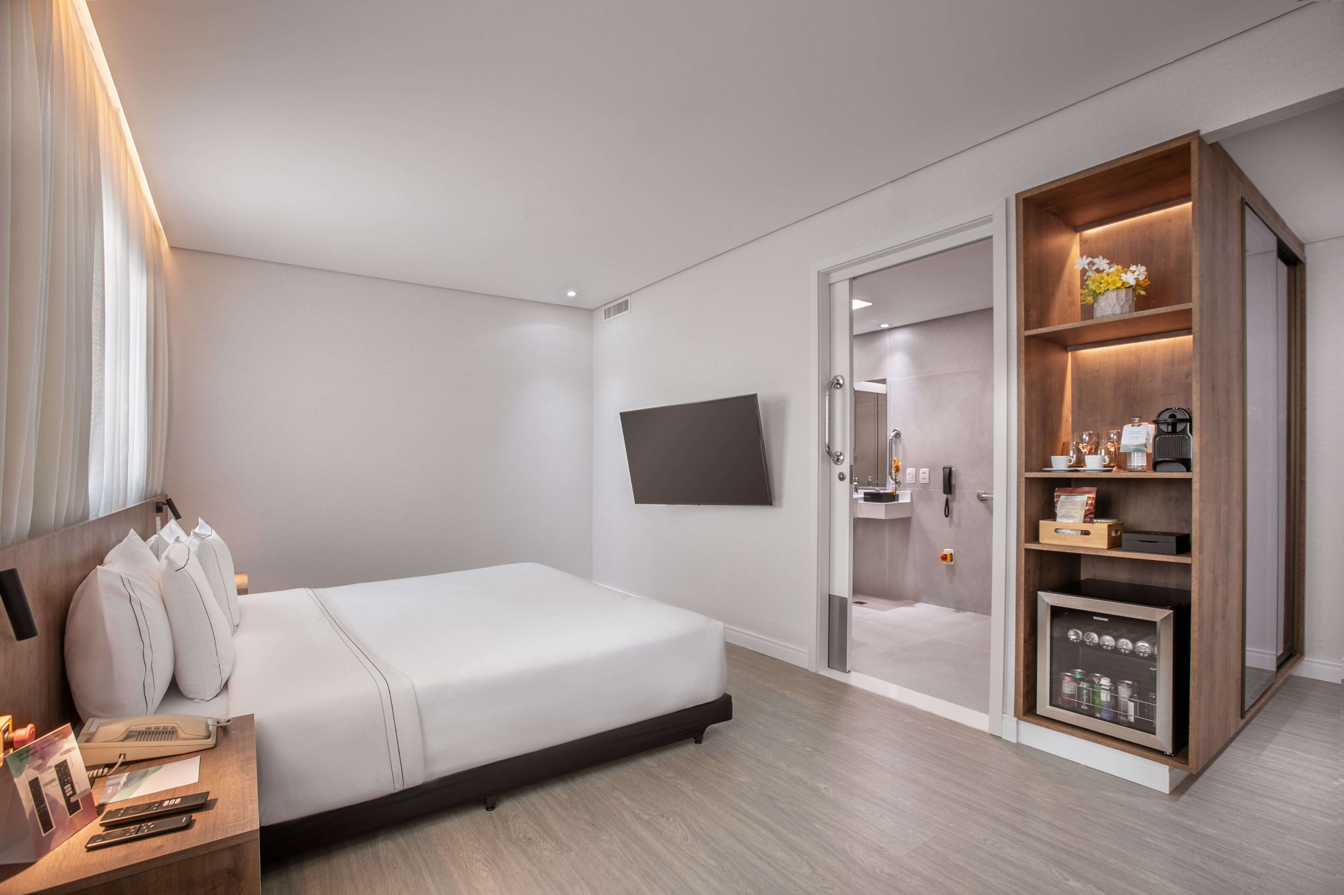 Minimalist hotel room with white bed, TV, minibar, and modern bathroom.