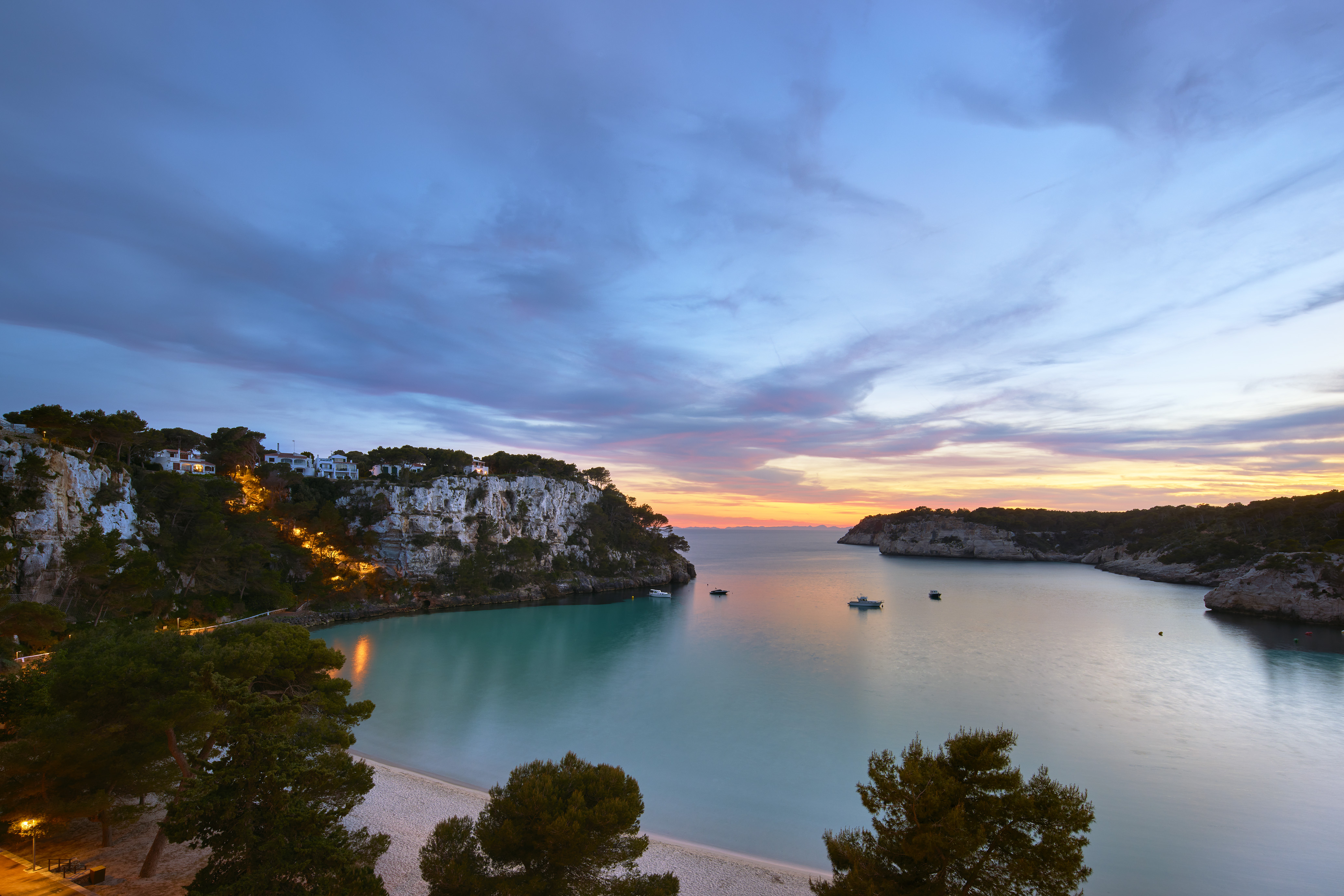 Turquoise bay at sunset with sandy beach, cliff houses, and boats.