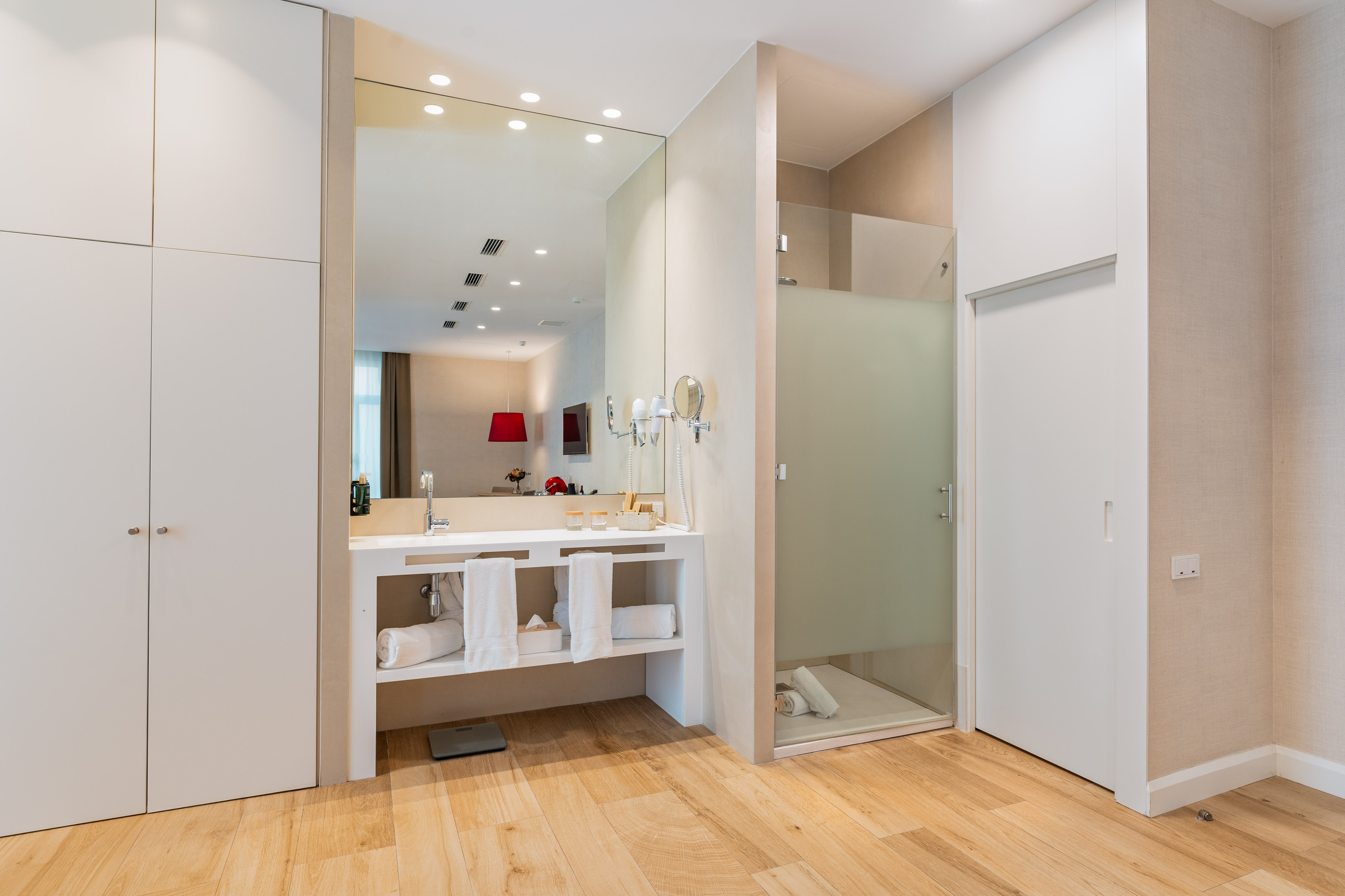 Modern hotel bathroom: white vanity, large mirror, wood floor, glass shower.