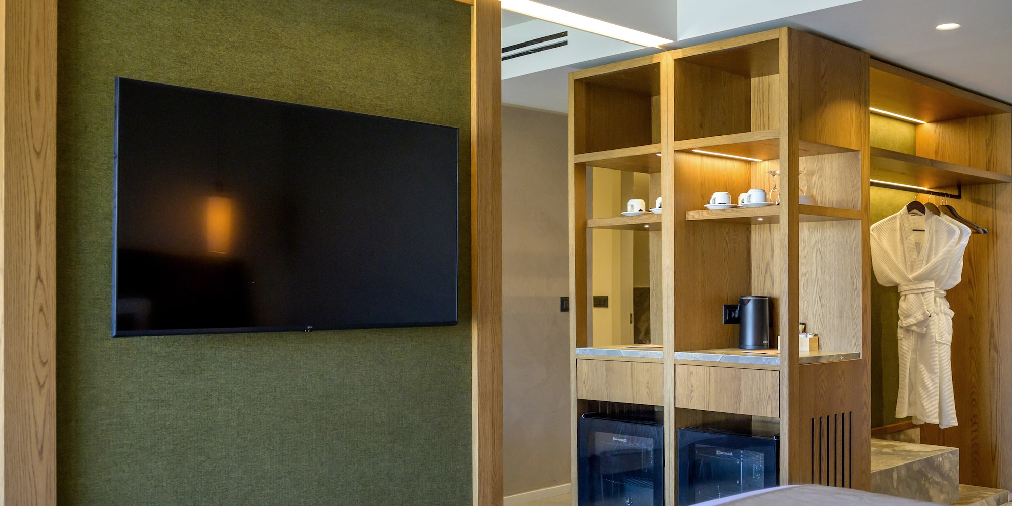 Modern hotel room with green wall, mounted TV, wooden cabinet, and minibar.