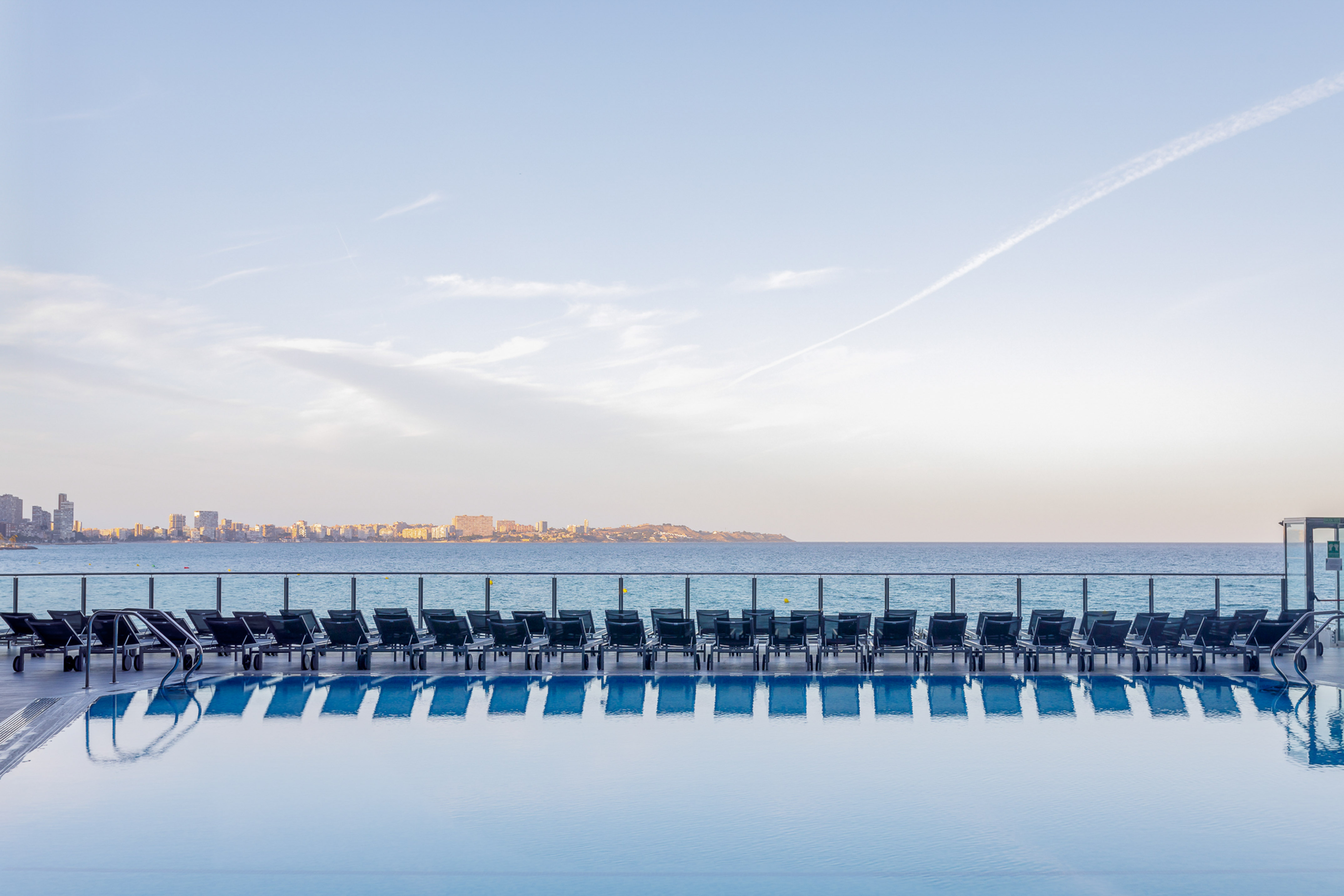 Infinity pool, sun loungers reflected in water, ocean, distant city skyline.