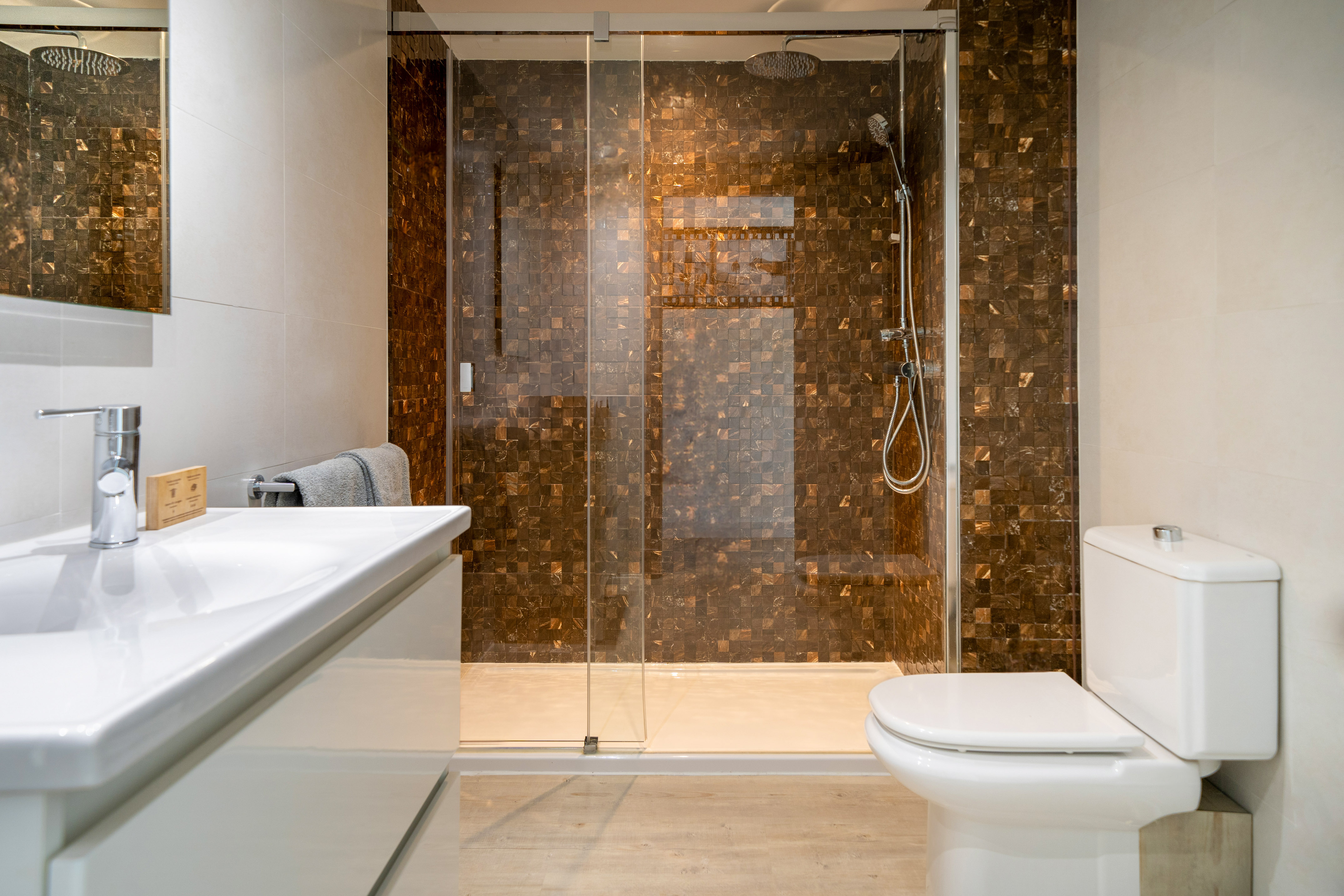 Modern bathroom with walk-in shower, brown mosaic tiles, white vanity, and toilet.