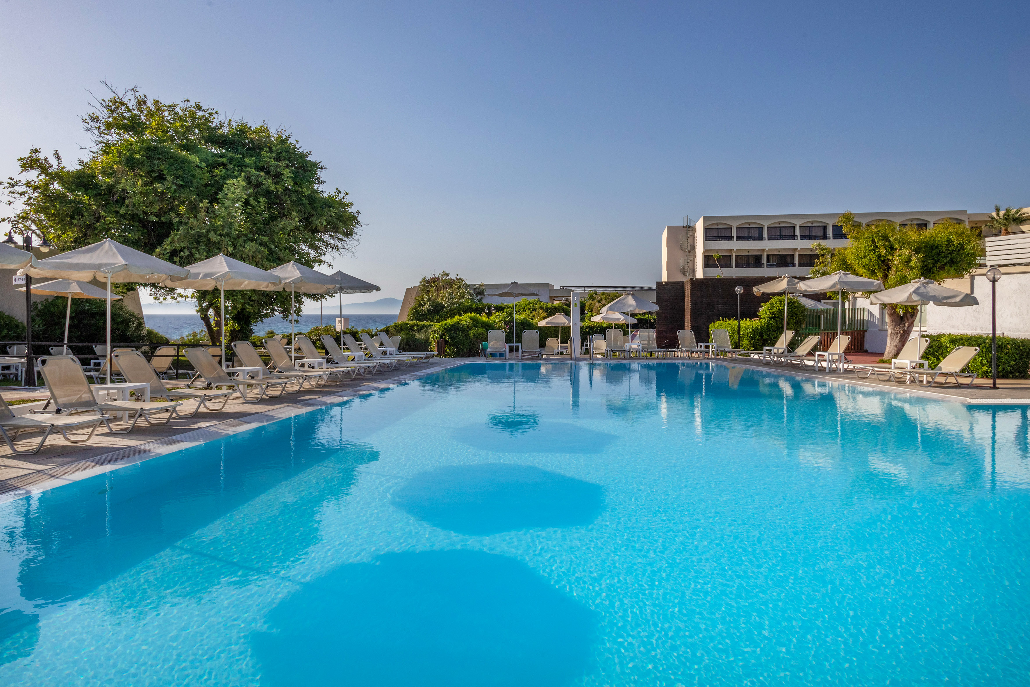 Sparkling blue hotel pool with lounge chairs, umbrellas, and distant ocean view.