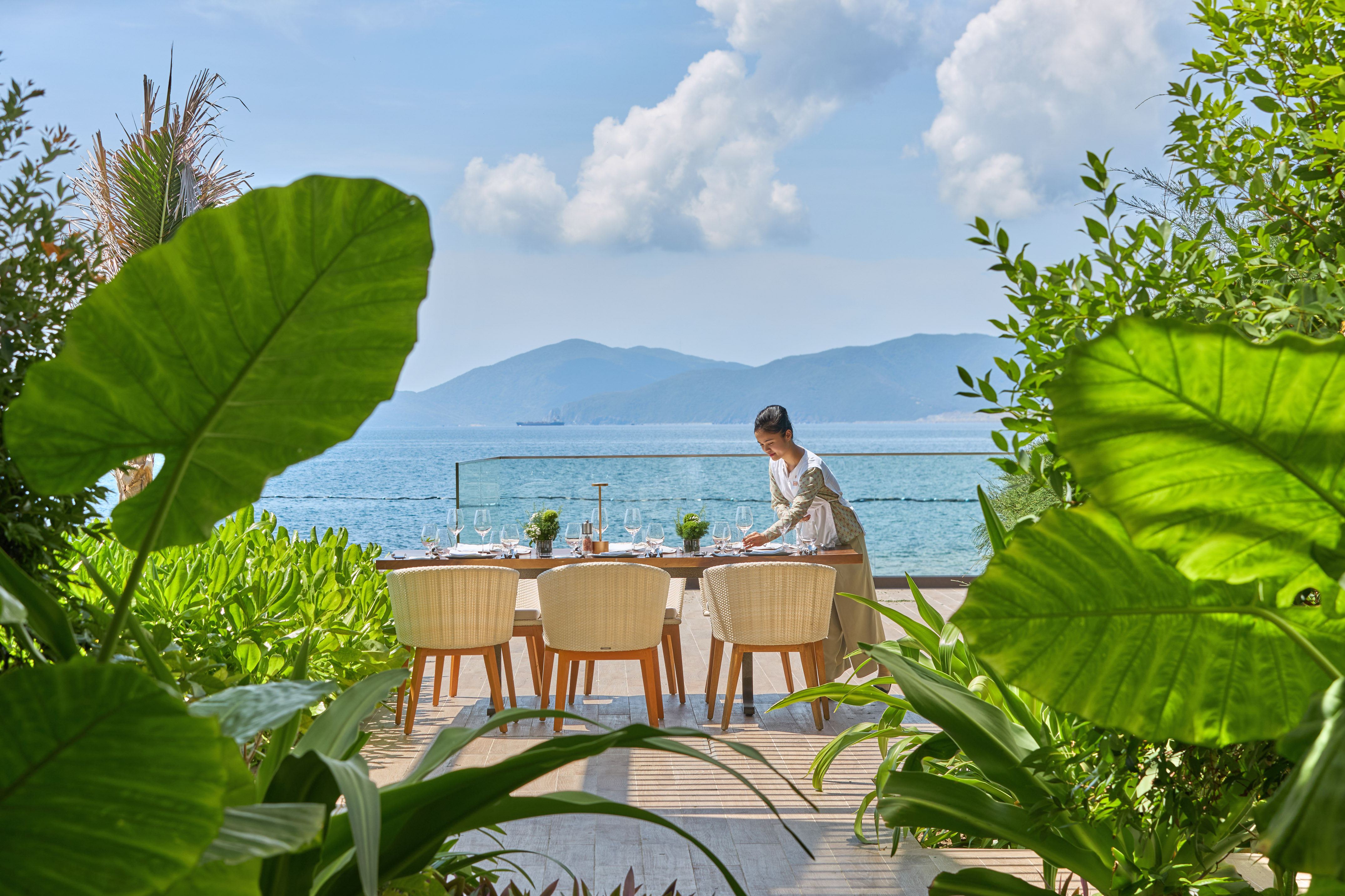 Staff prepares outdoor dining table with ocean view and tropical plants.