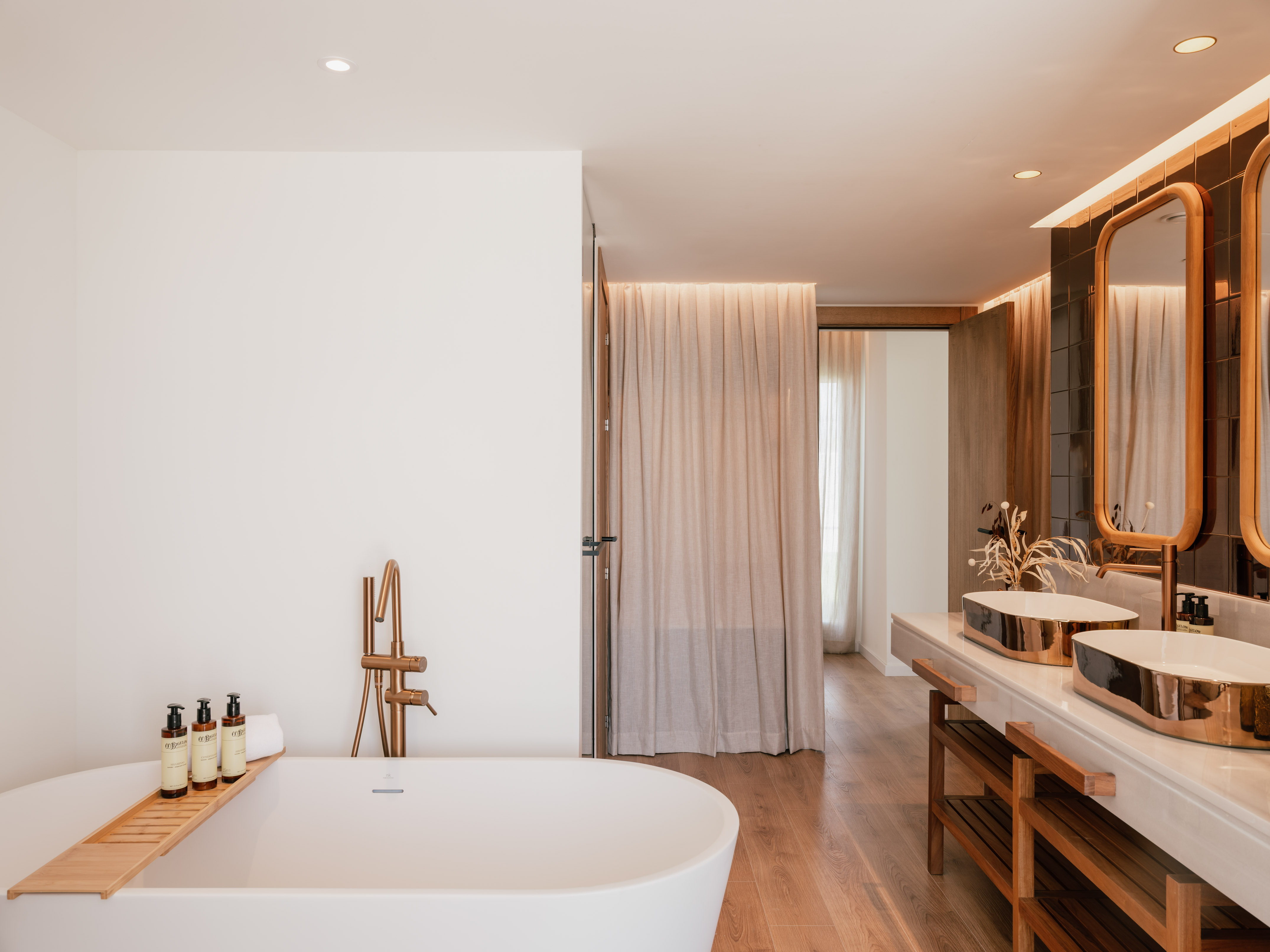 Modern luxury bathroom with white tub, copper faucet, dual vessel sinks, wood floor.