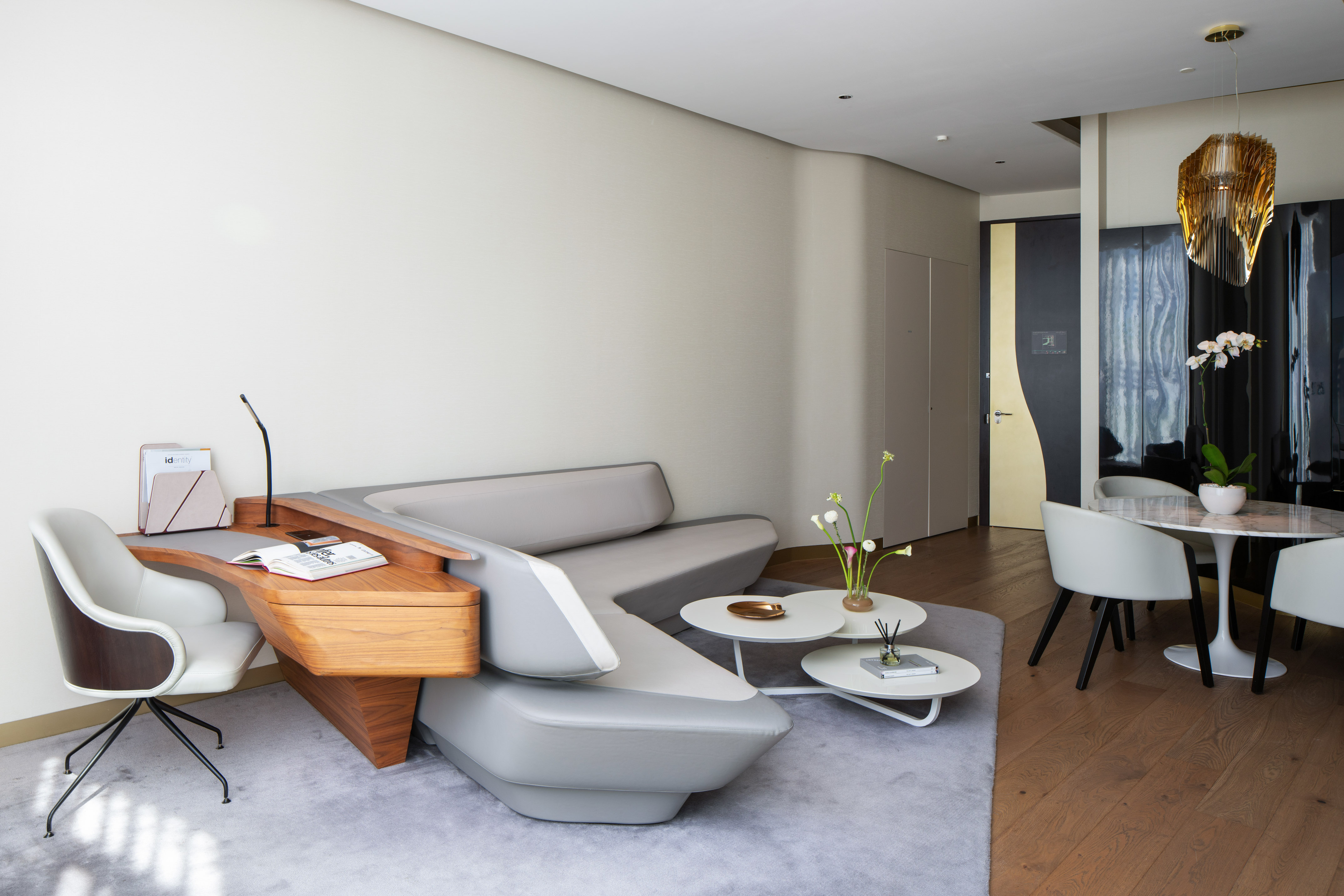 Luxury hotel suite with modern gray sofa, wooden desk, and contemporary dining.