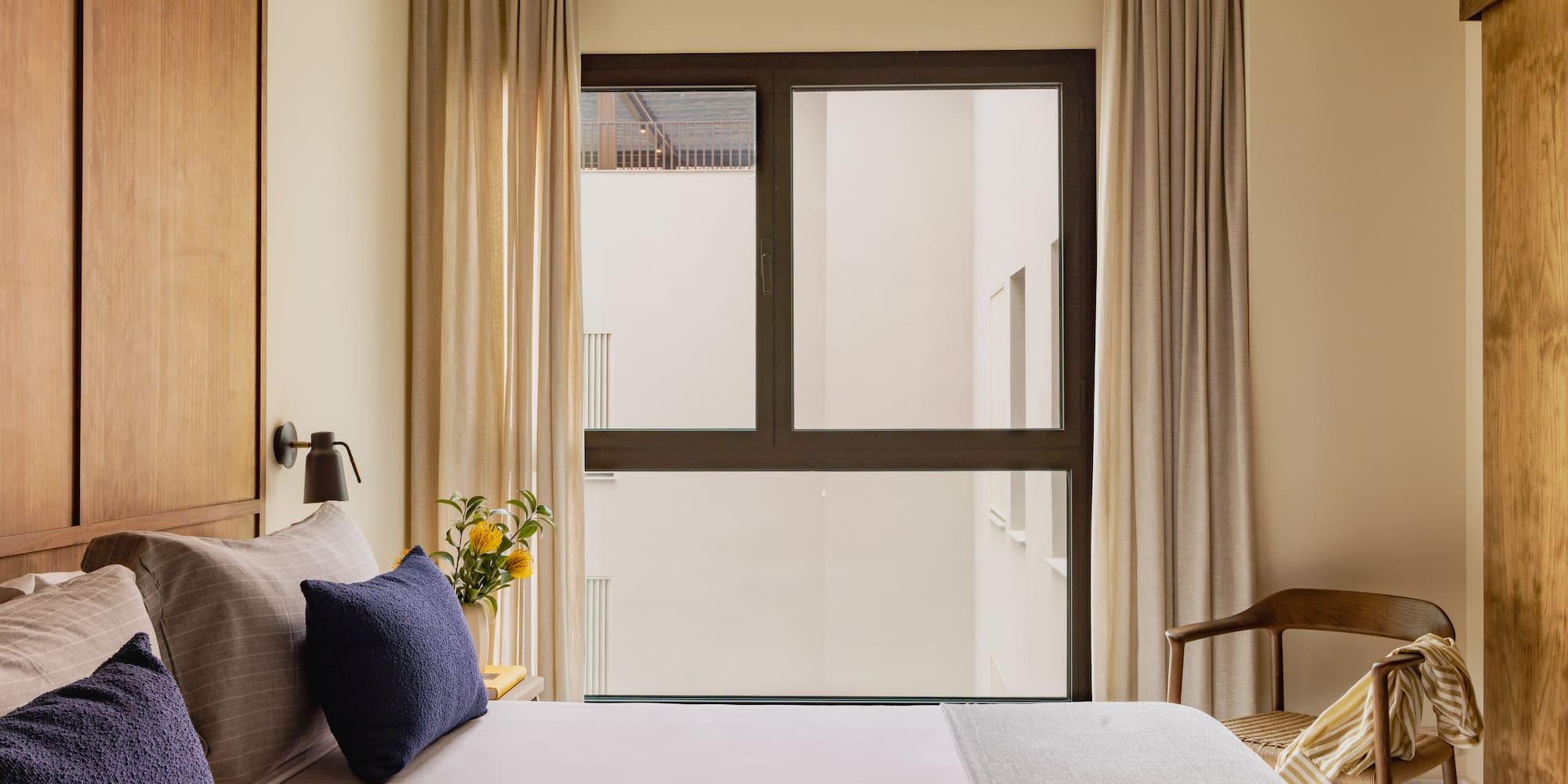 Modern hotel bedroom with wooden headboard, navy pillows, and large window.