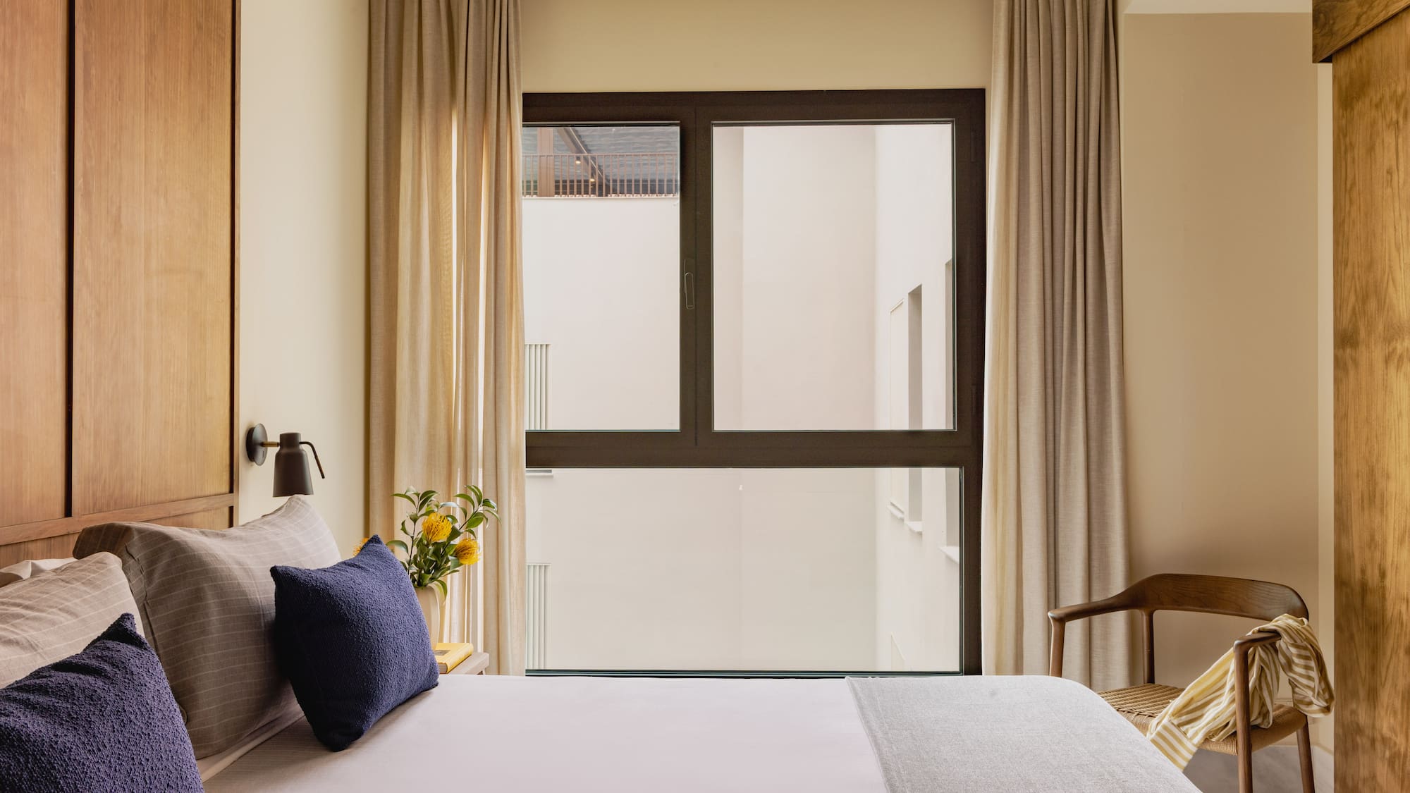 Modern hotel bedroom with wooden headboard, navy pillows, and large window.