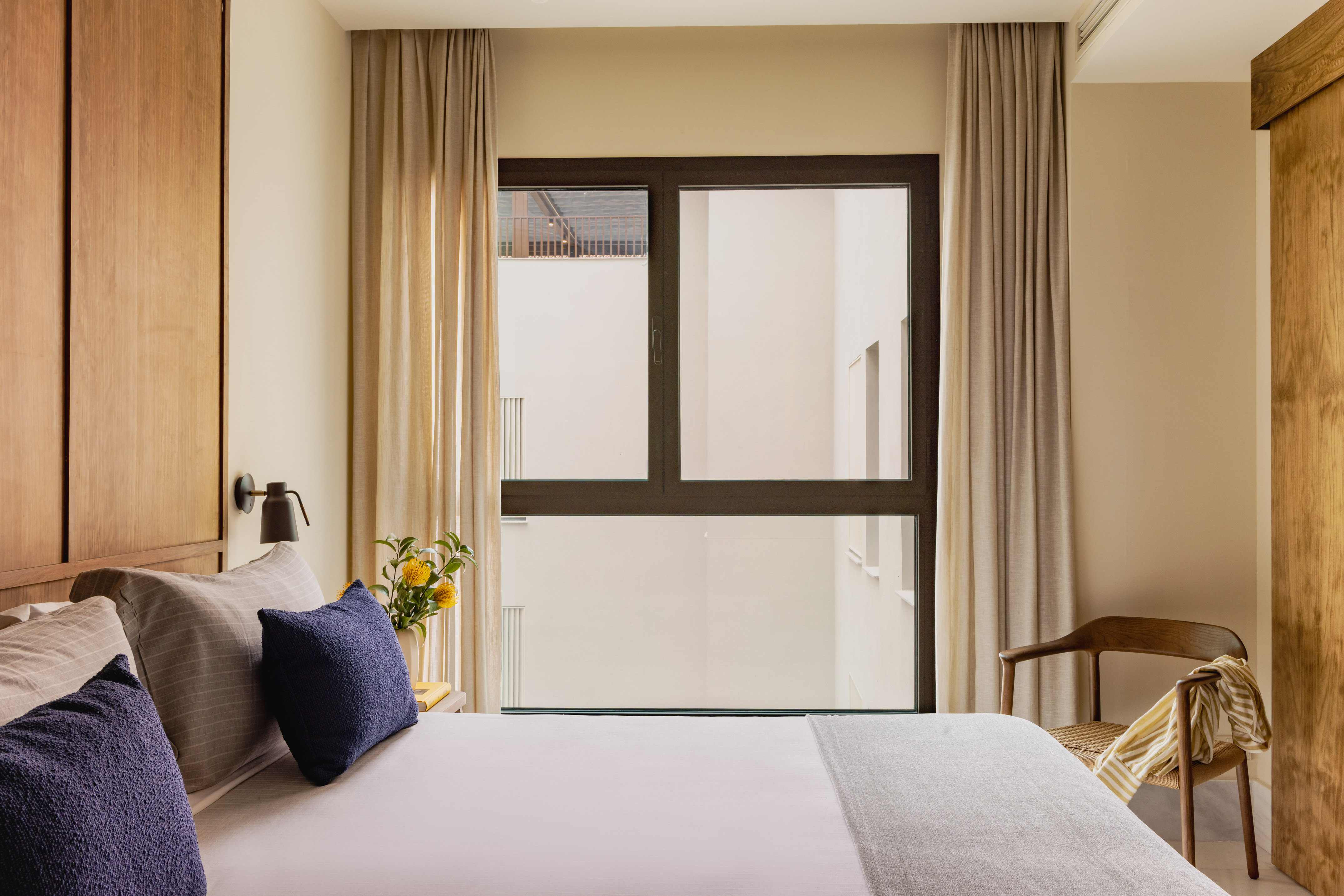 Modern hotel bedroom with wooden headboard, navy pillows, and large window.