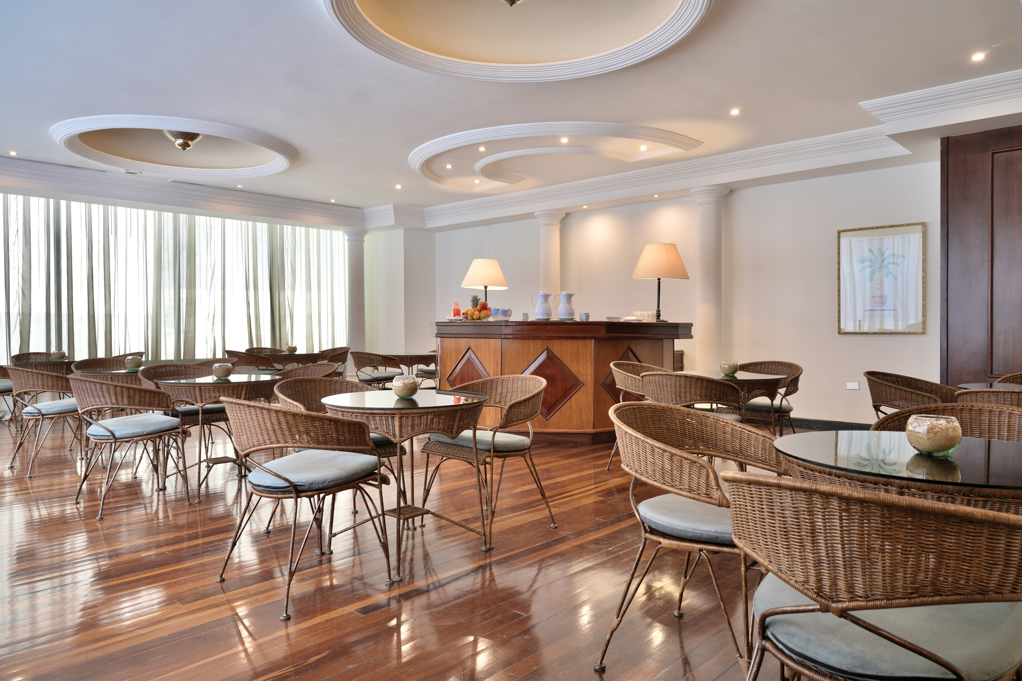 Bright cafe lounge with wicker chairs, glass tables, and a wooden bar.