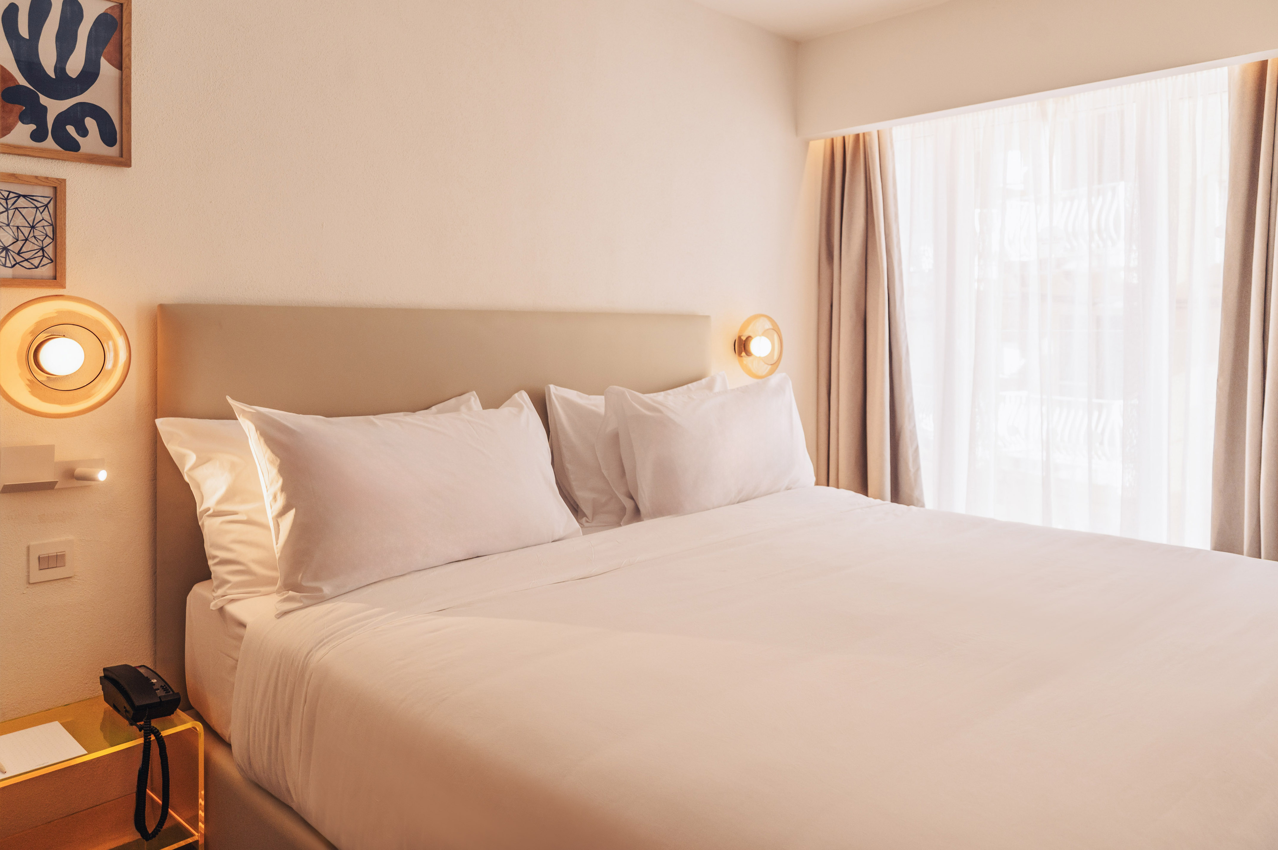 Bright, modern hotel room with white bed, beige headboard, sconces, and window.