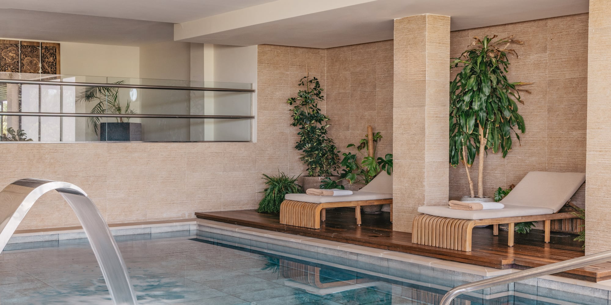 Indoor spa pool with waterfall, wooden loungers, and tropical plants.