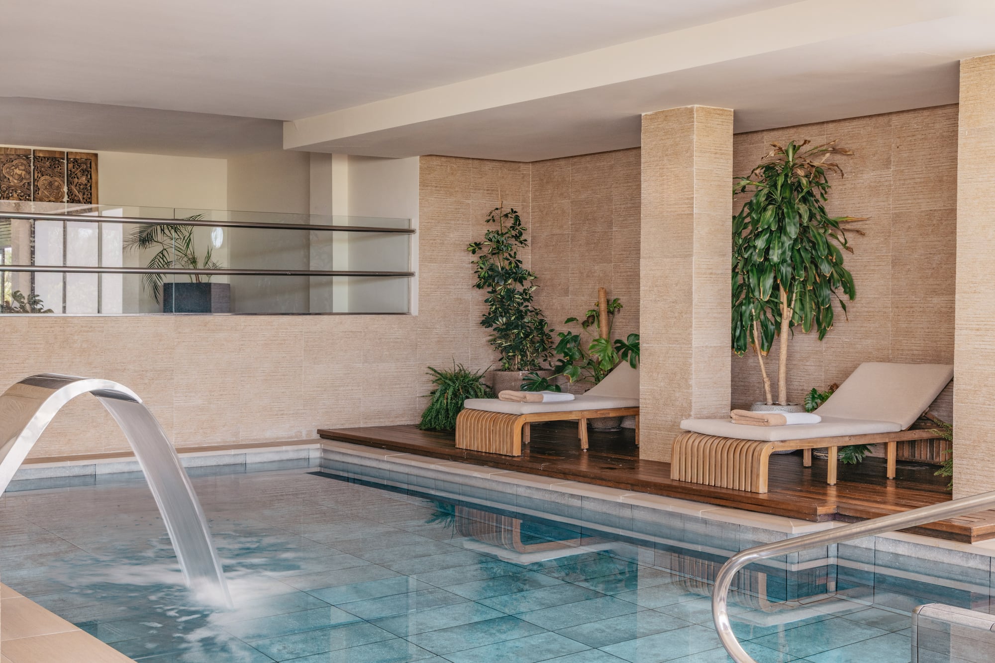 Indoor spa pool with waterfall, lounge chairs, and tropical plants.