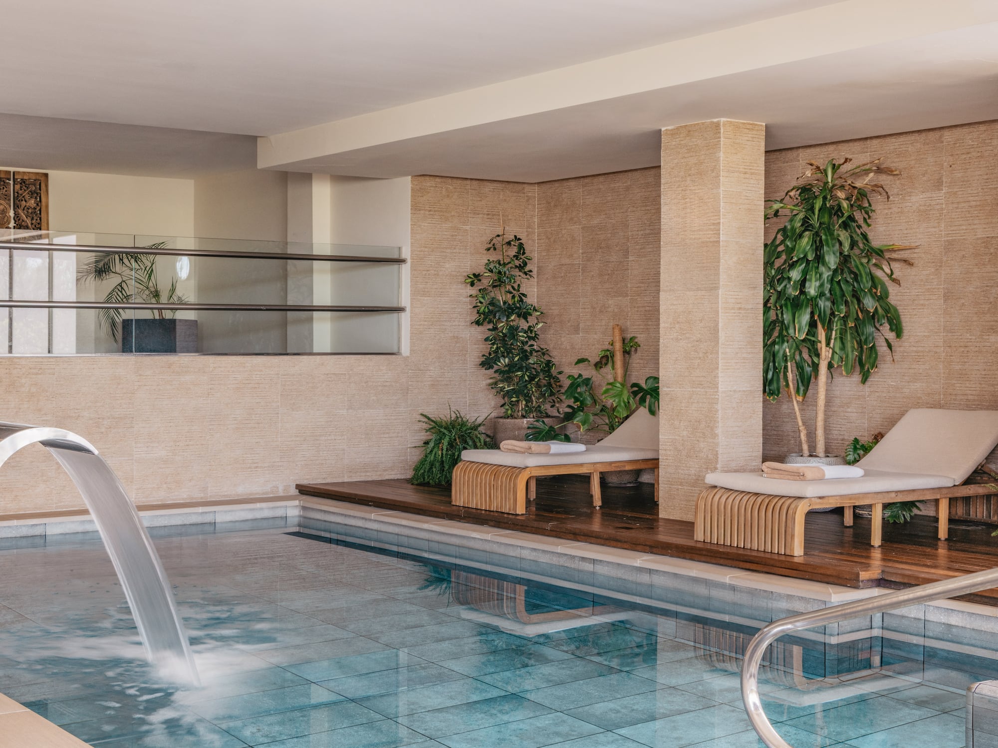 Indoor spa pool with waterfall, lounge chairs, and tropical plants.