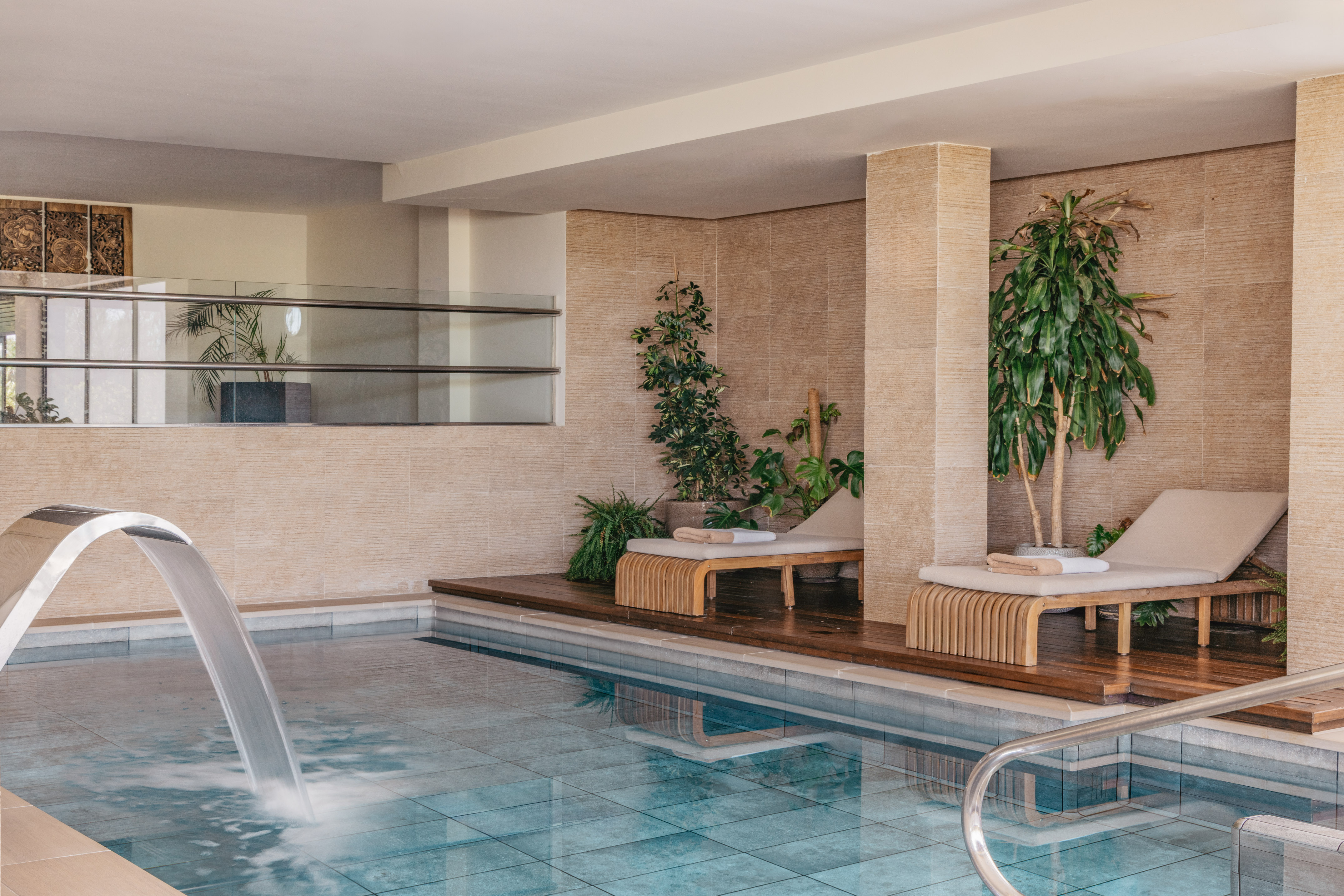 Indoor spa pool with waterfall, lounge chairs, and tropical plants.