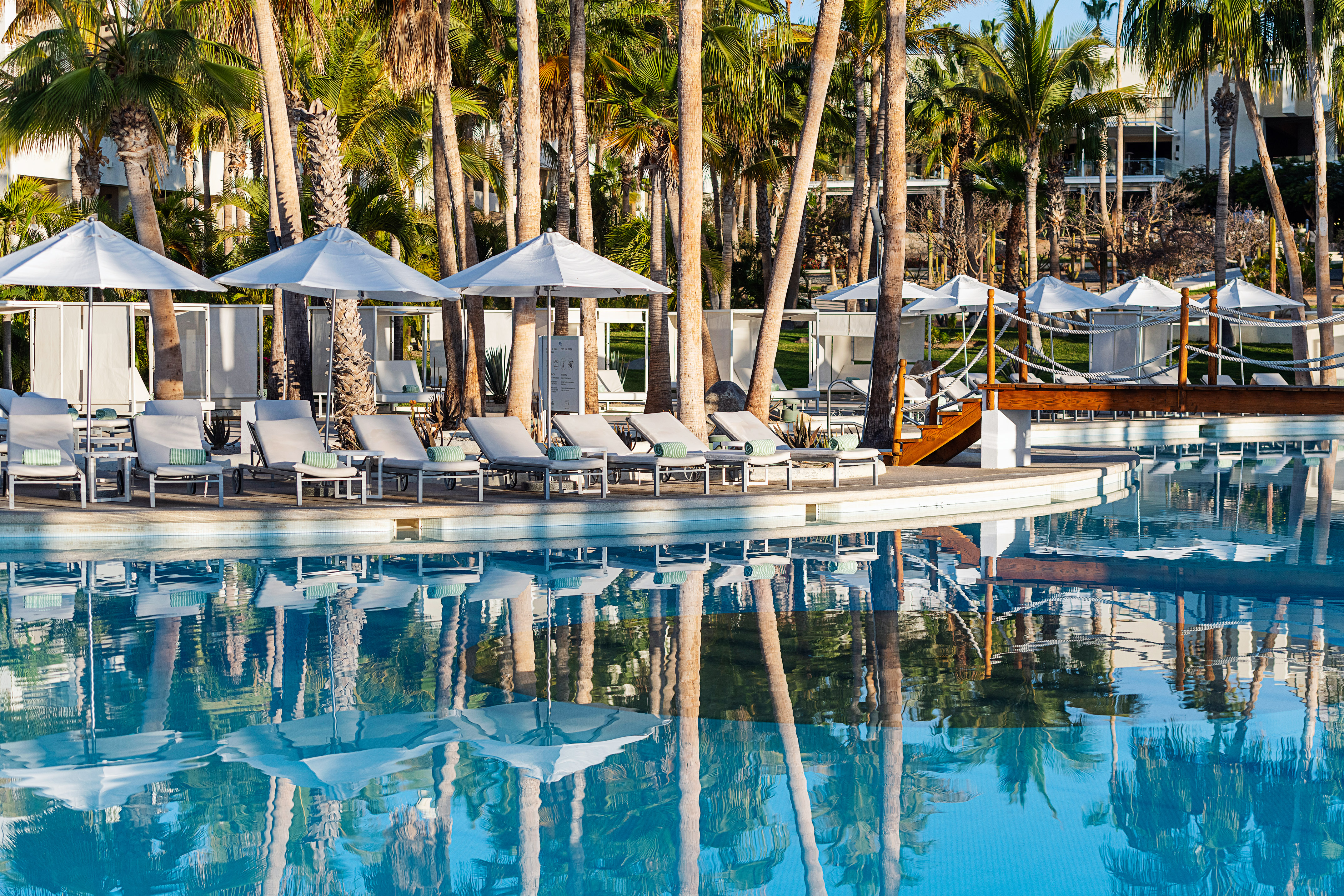 Resort pool with lounge chairs, umbrellas, and palm trees reflected in water.