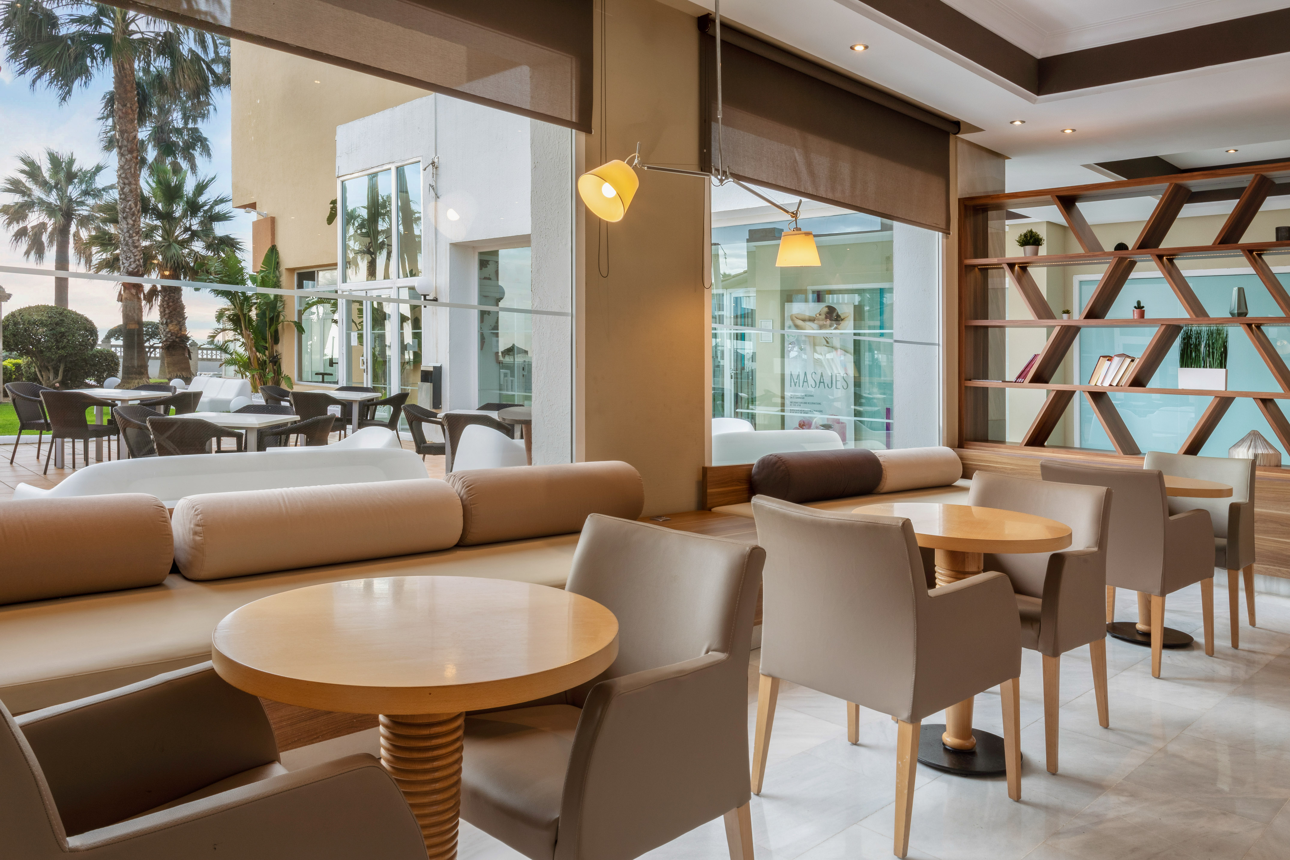 Modern resort lounge with cozy seating, wooden tables, and palm tree view.