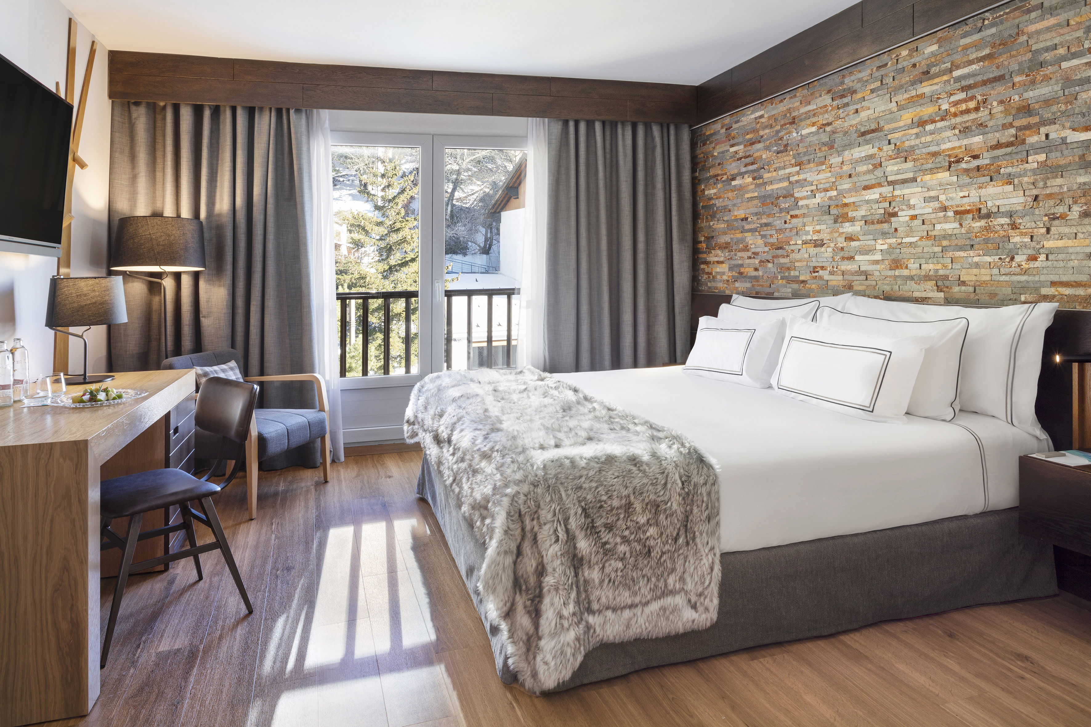 Luxury hotel bedroom with stone wall, king bed, desk, and snowy mountain view.