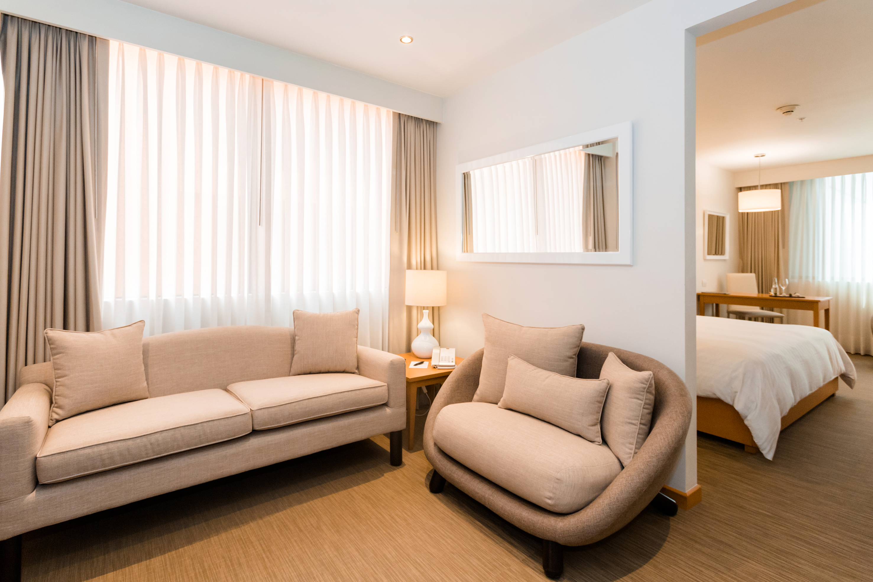 Modern hotel suite living room with beige sofa, armchair, and bedroom.