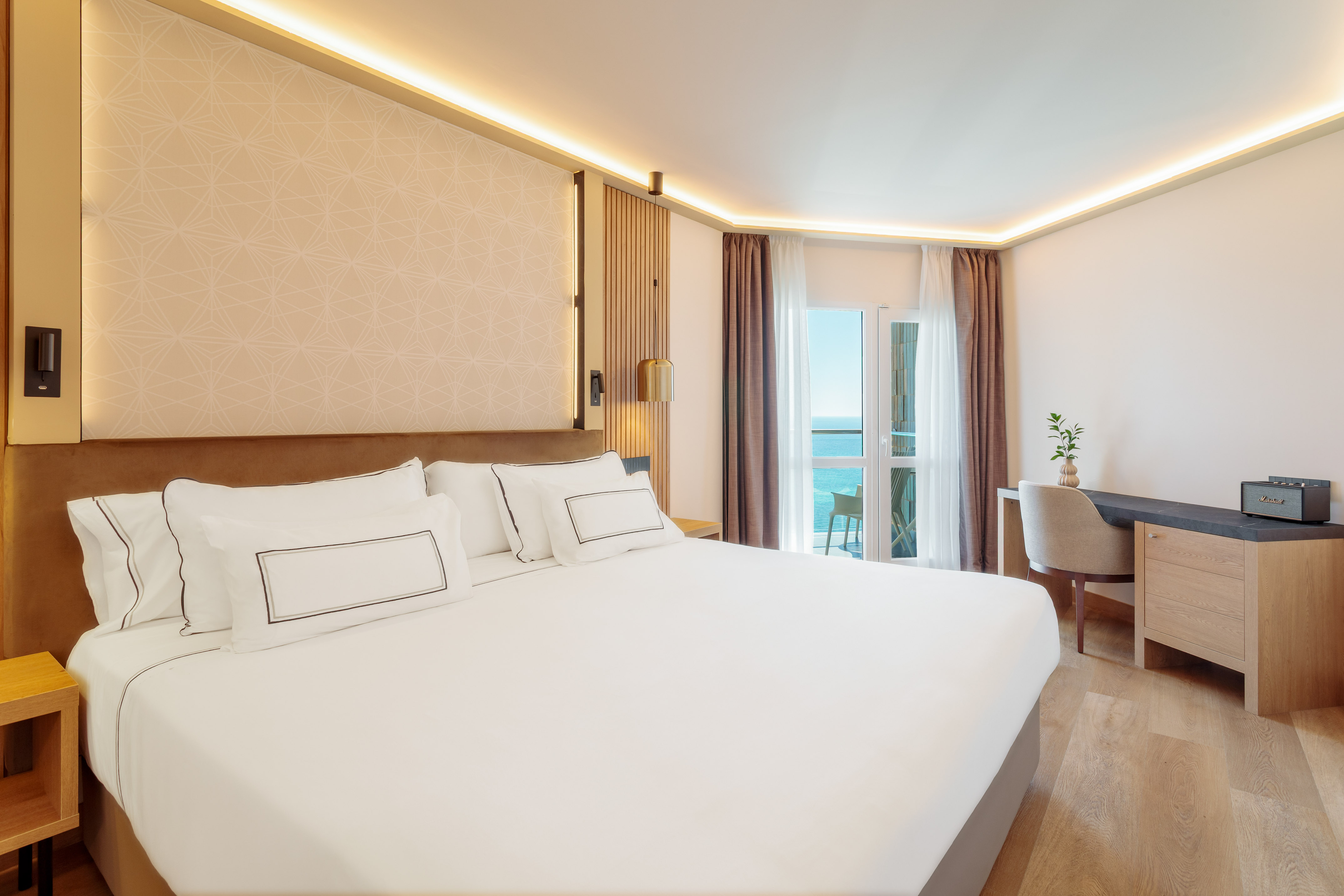 Modern hotel bedroom with white bed, desk, and balcony overlooking the sea.