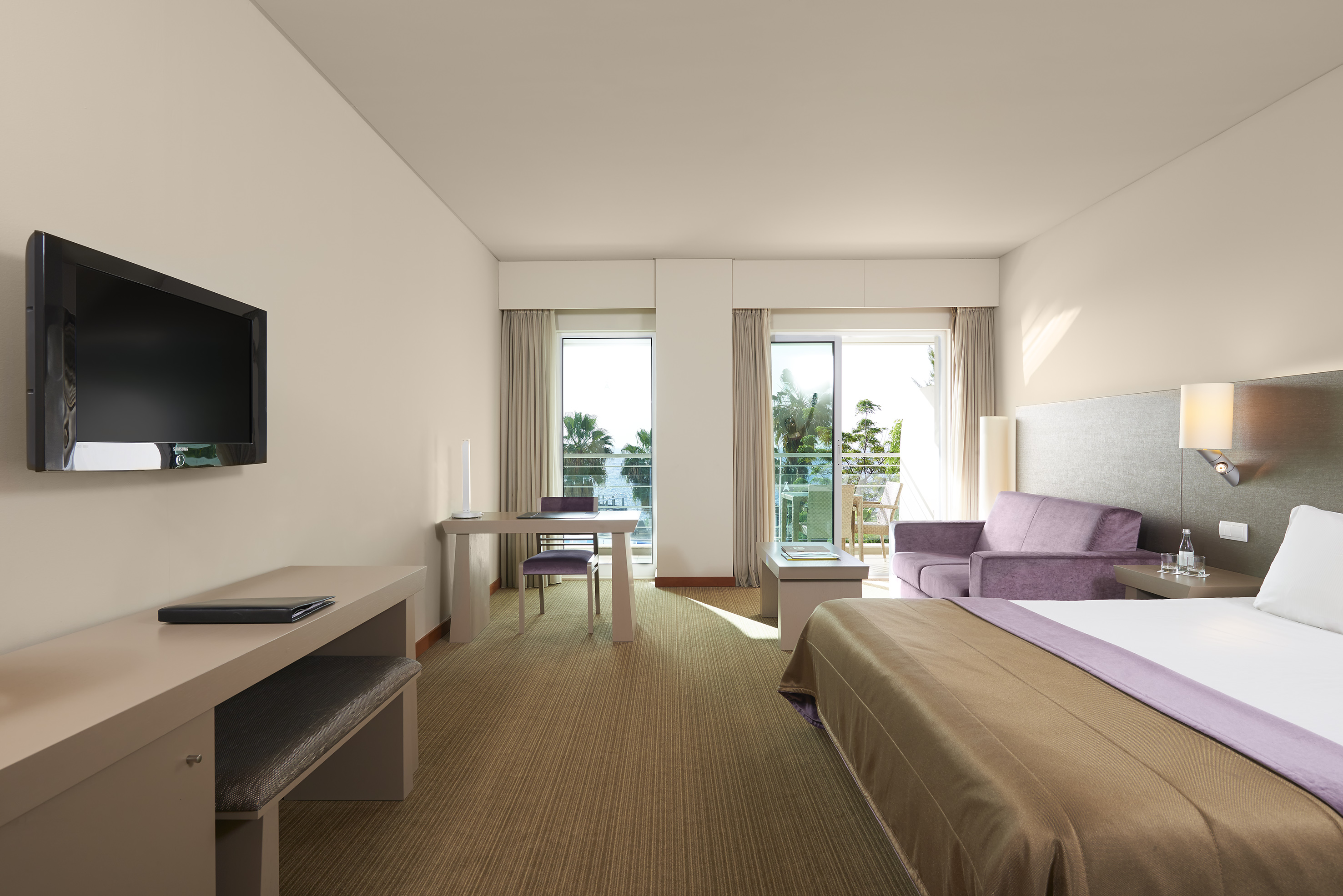 Modern hotel suite with bed, purple sofa, desk, TV, and balcony view of palm trees.