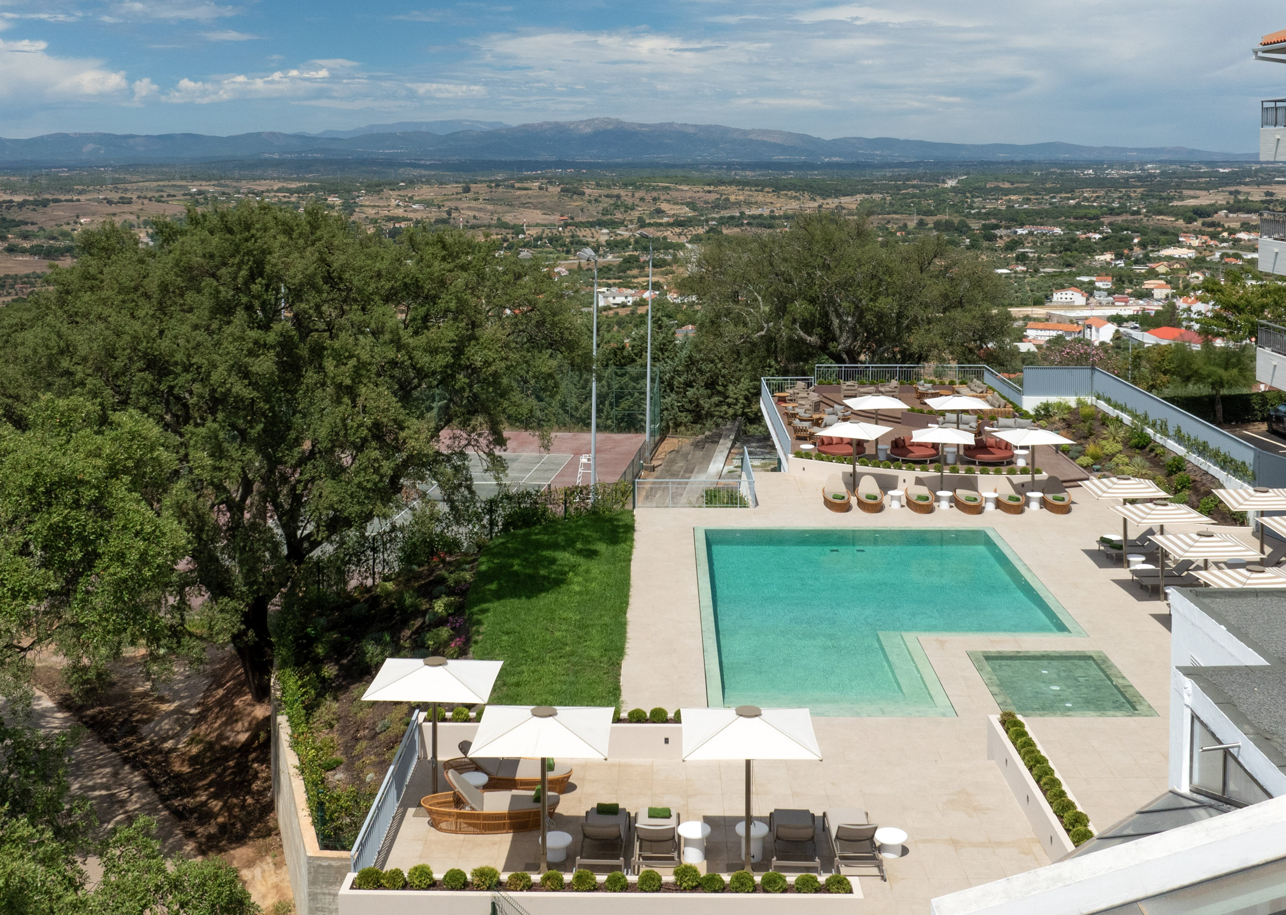 Aerial resort view: turquoise pool, lounge, tennis court, distant mountains.