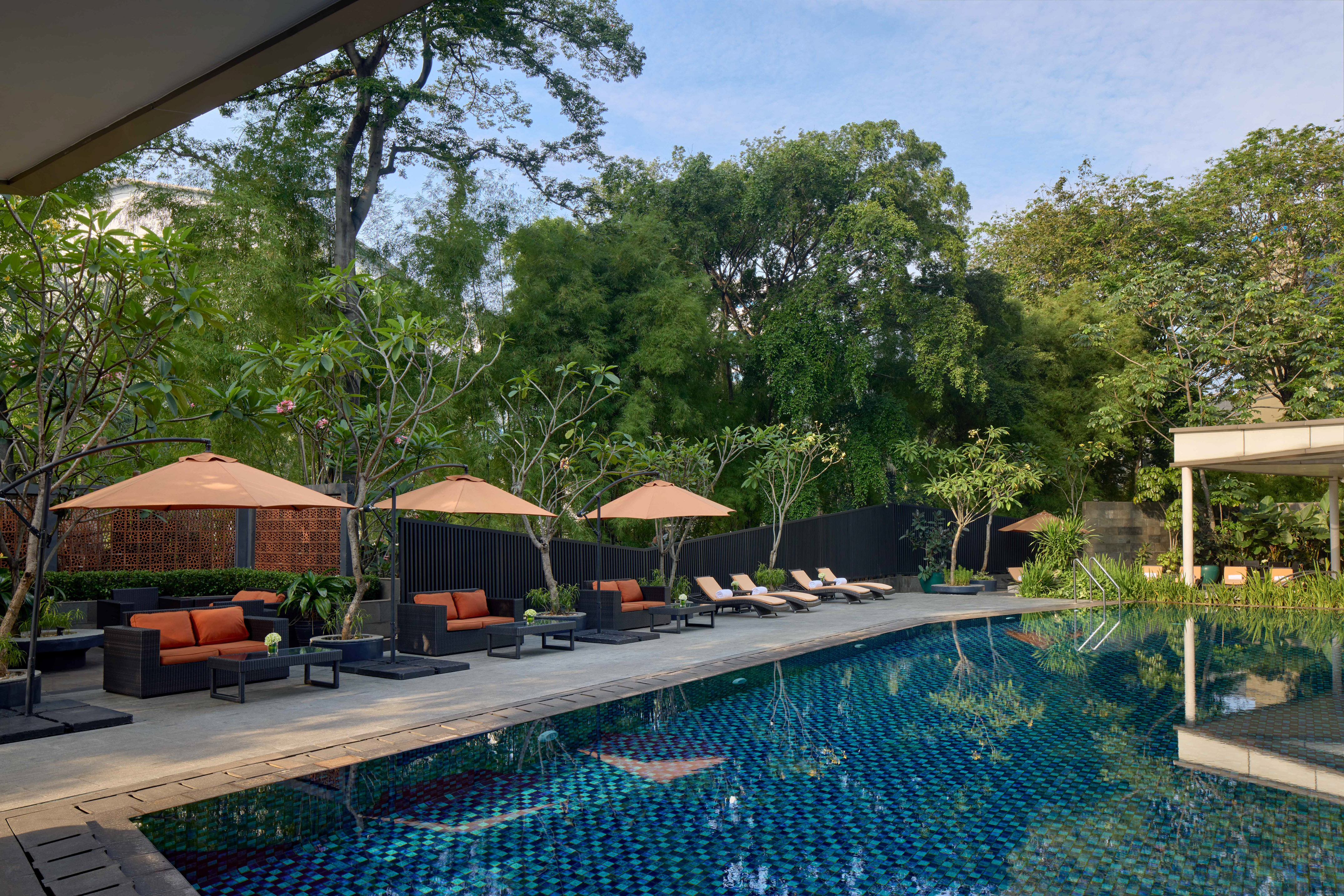 Outdoor resort pool with lounge chairs, orange sofas, umbrellas, and tropical trees.