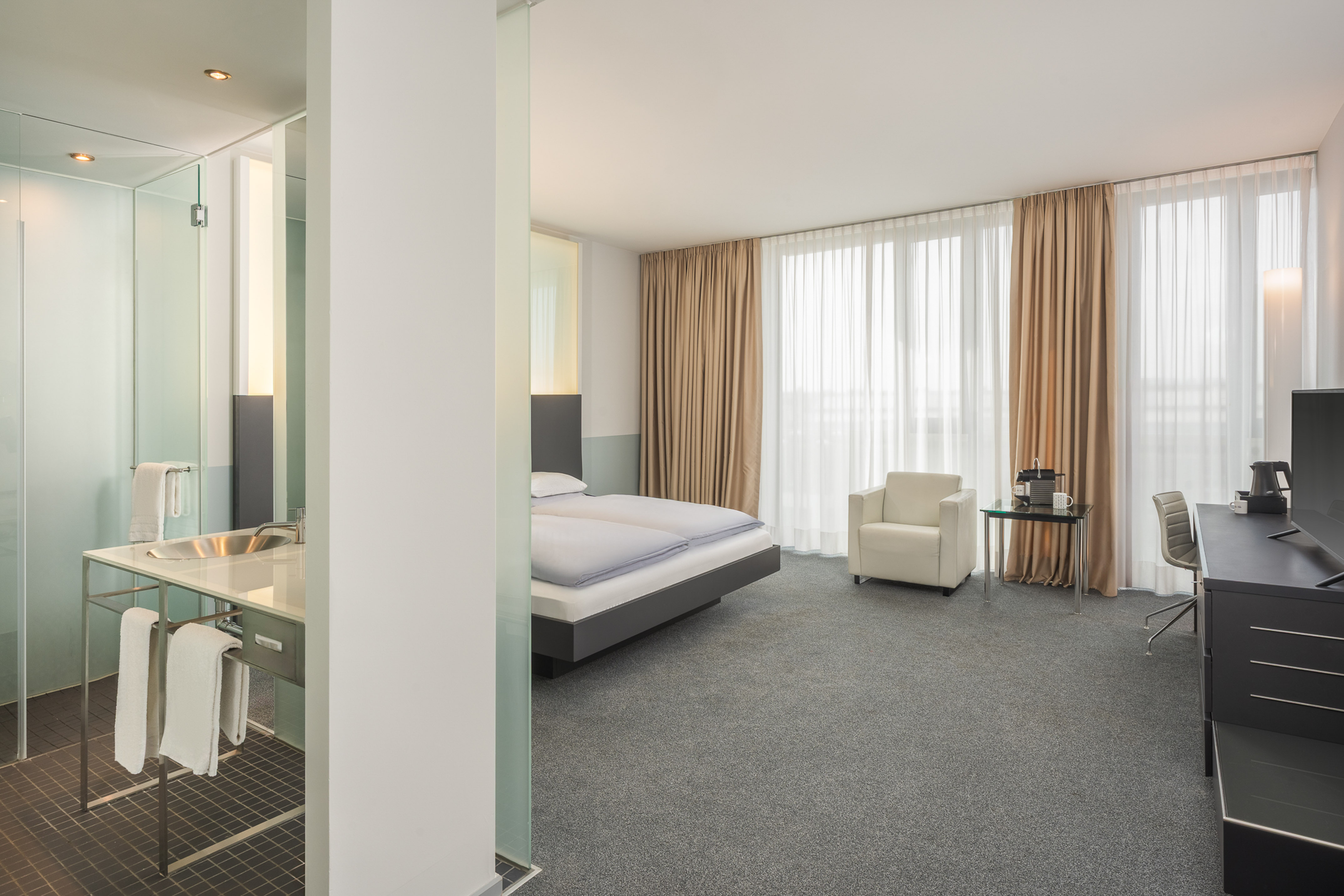 Modern hotel room with bed, work desk, armchair, and open-plan bathroom.