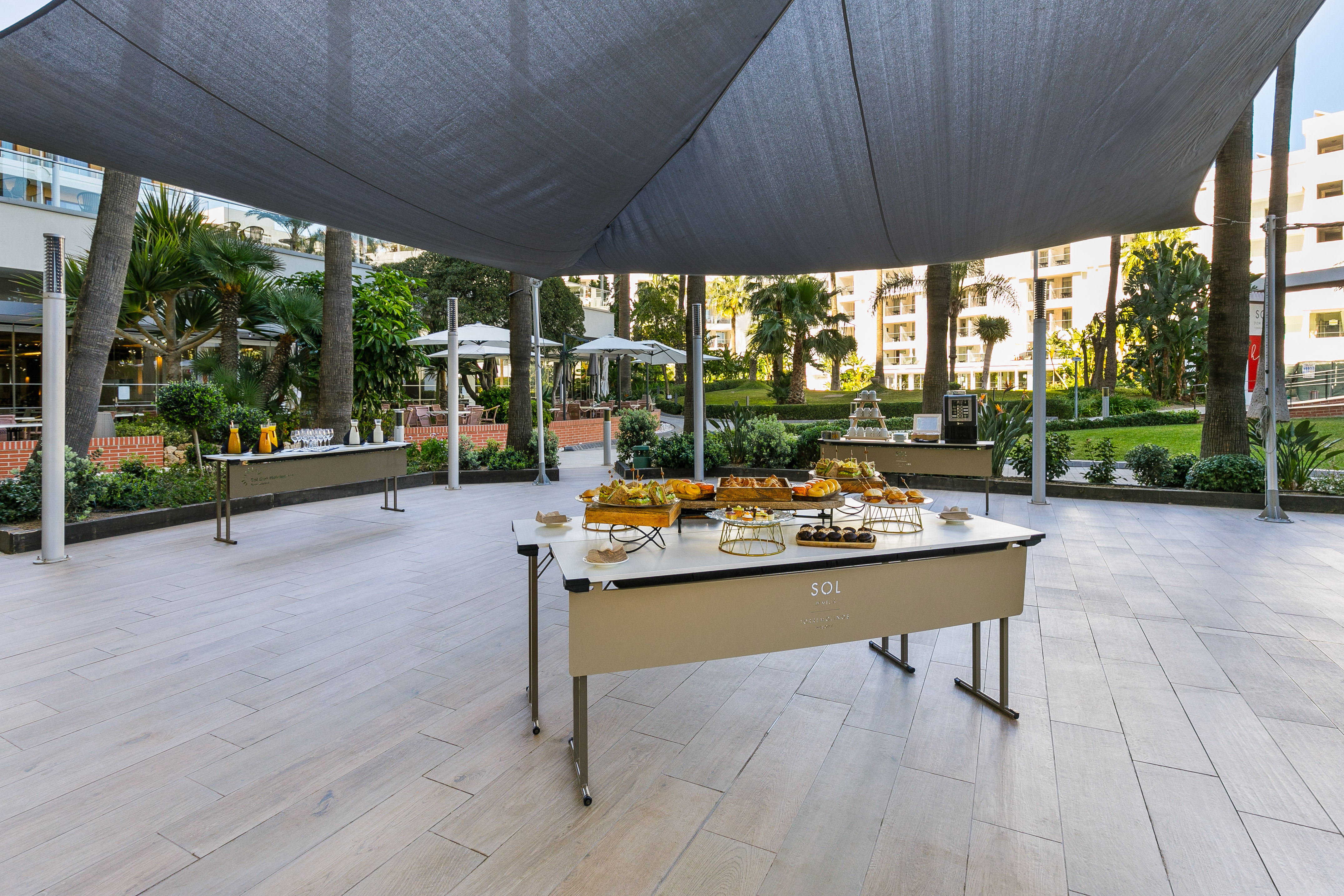 Outdoor hotel breakfast buffet on shaded terrace with food and palm trees.