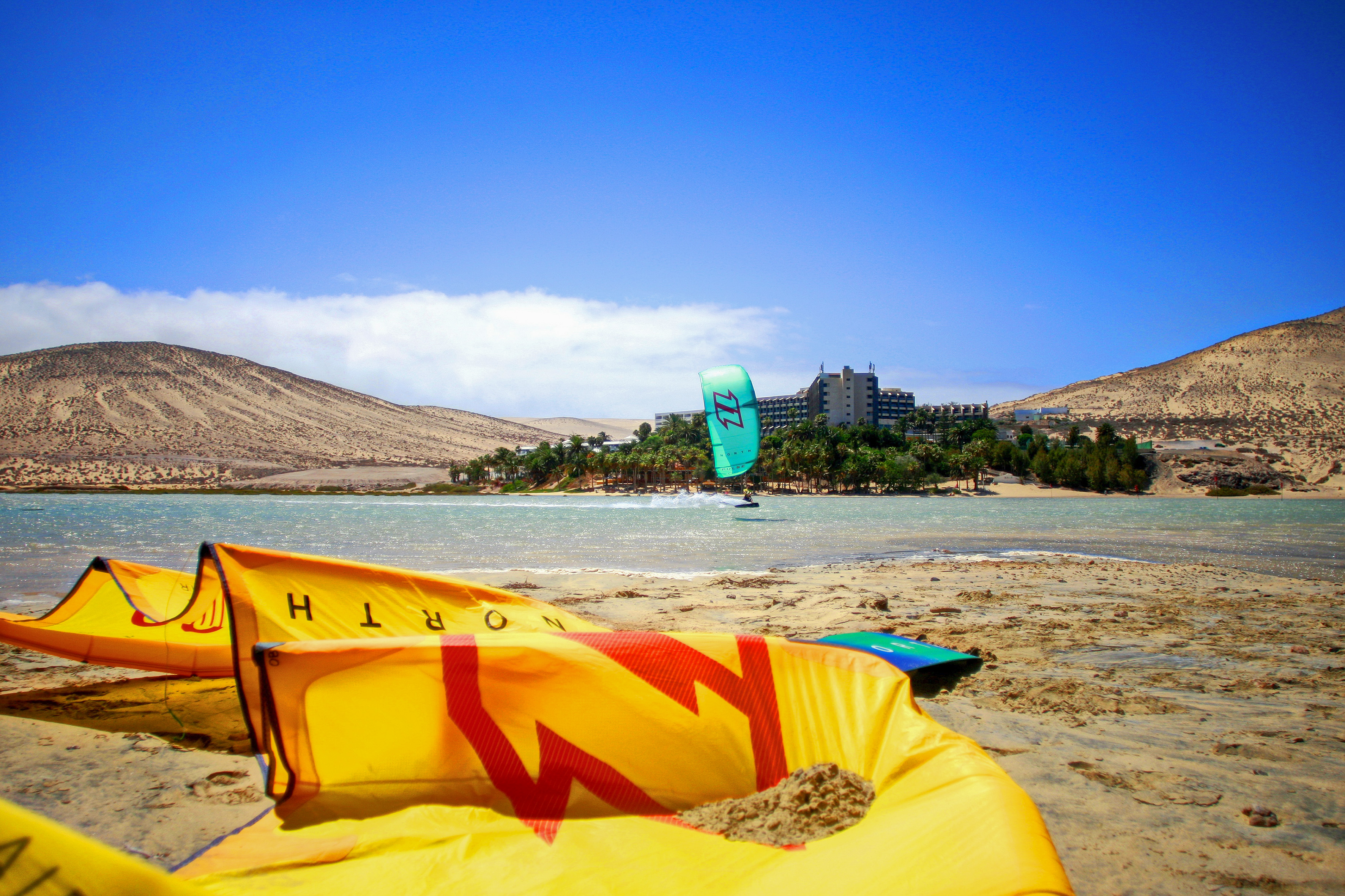 Kitesurfing lagoon with yellow kites on sandy beach, hotel resort, and dunes.