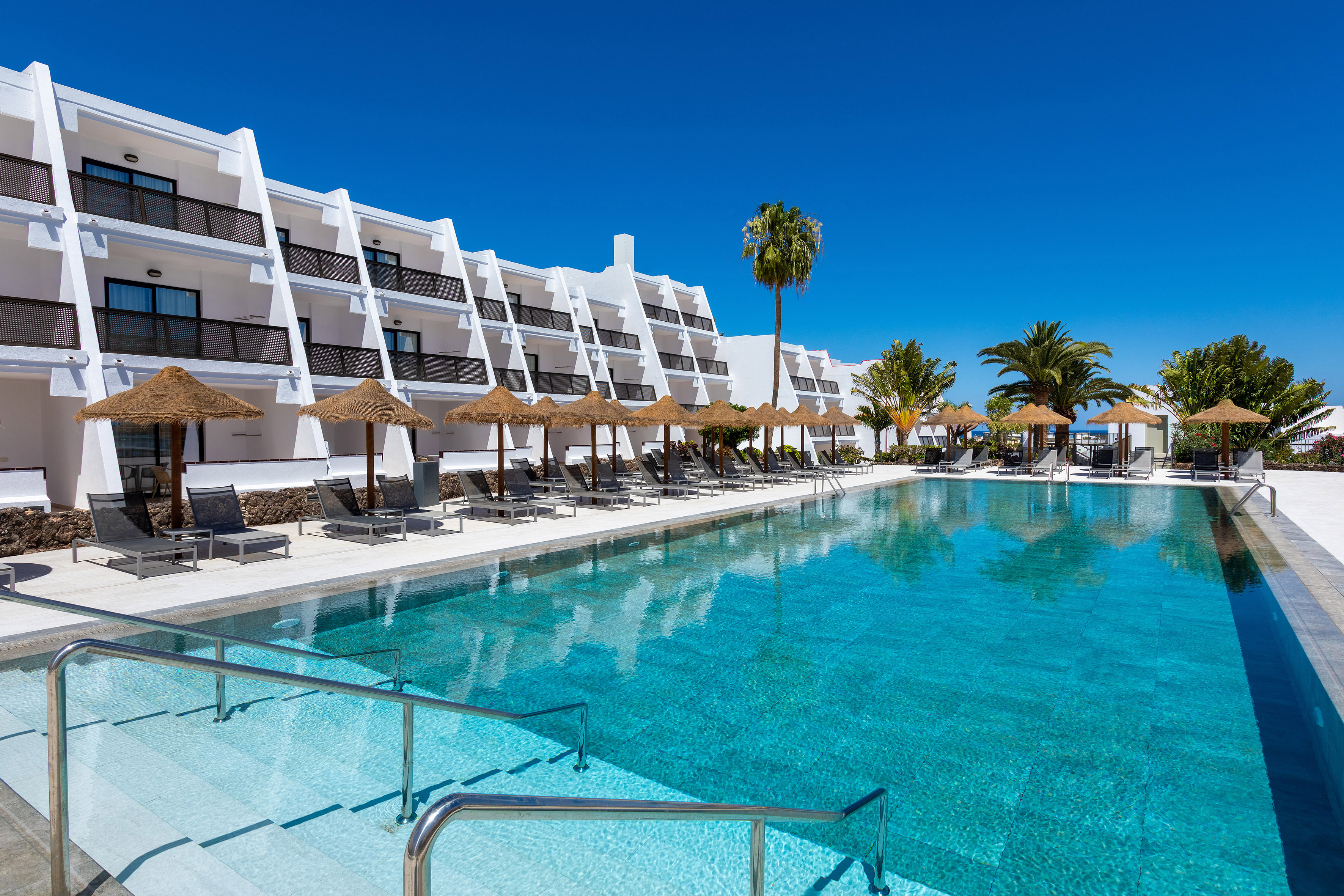 Sunny hotel resort pool with white buildings, palm trees, and lounge chairs.