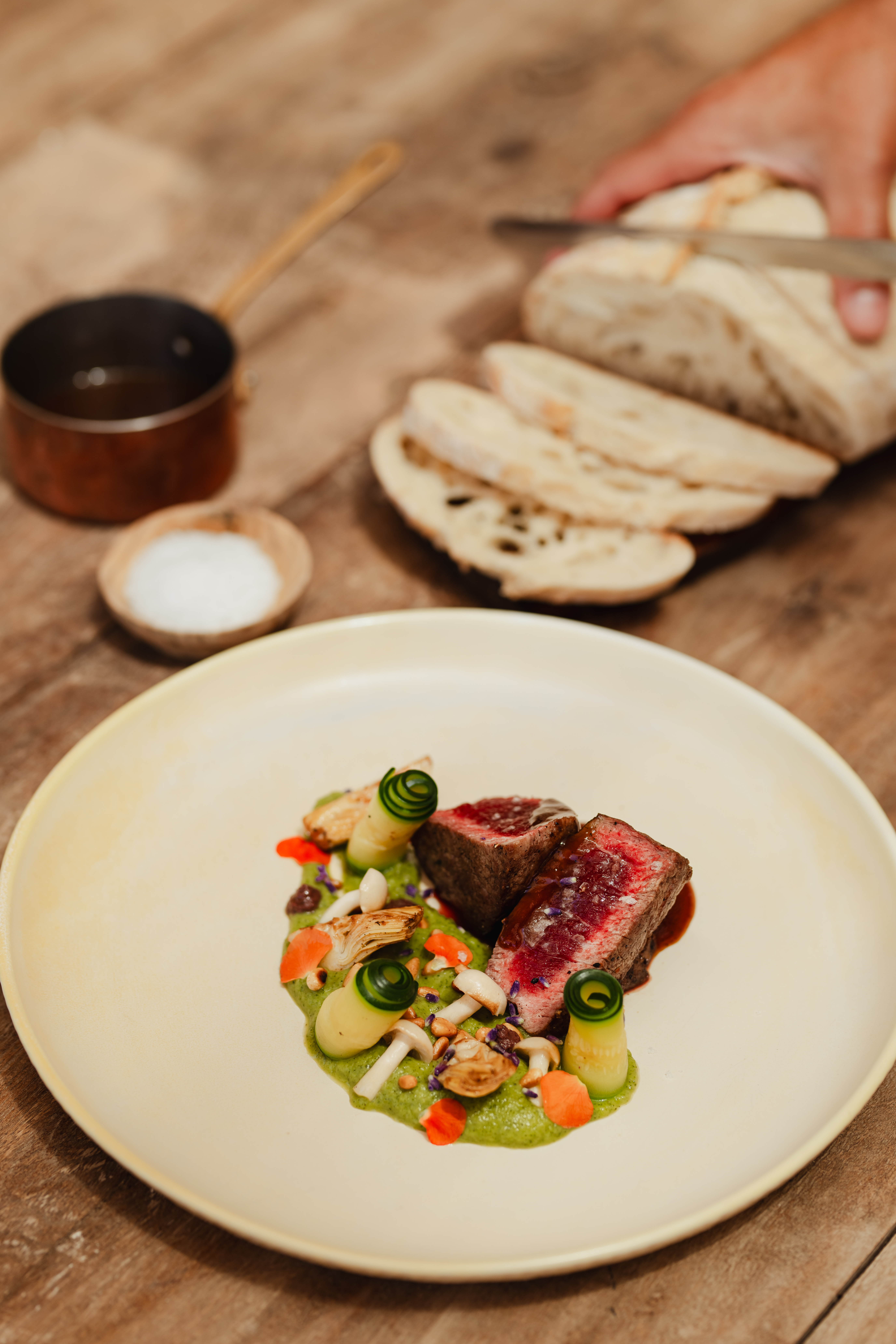Plated medium-rare steak with green puree, mushrooms, cucumber. Sliced bread, salt.