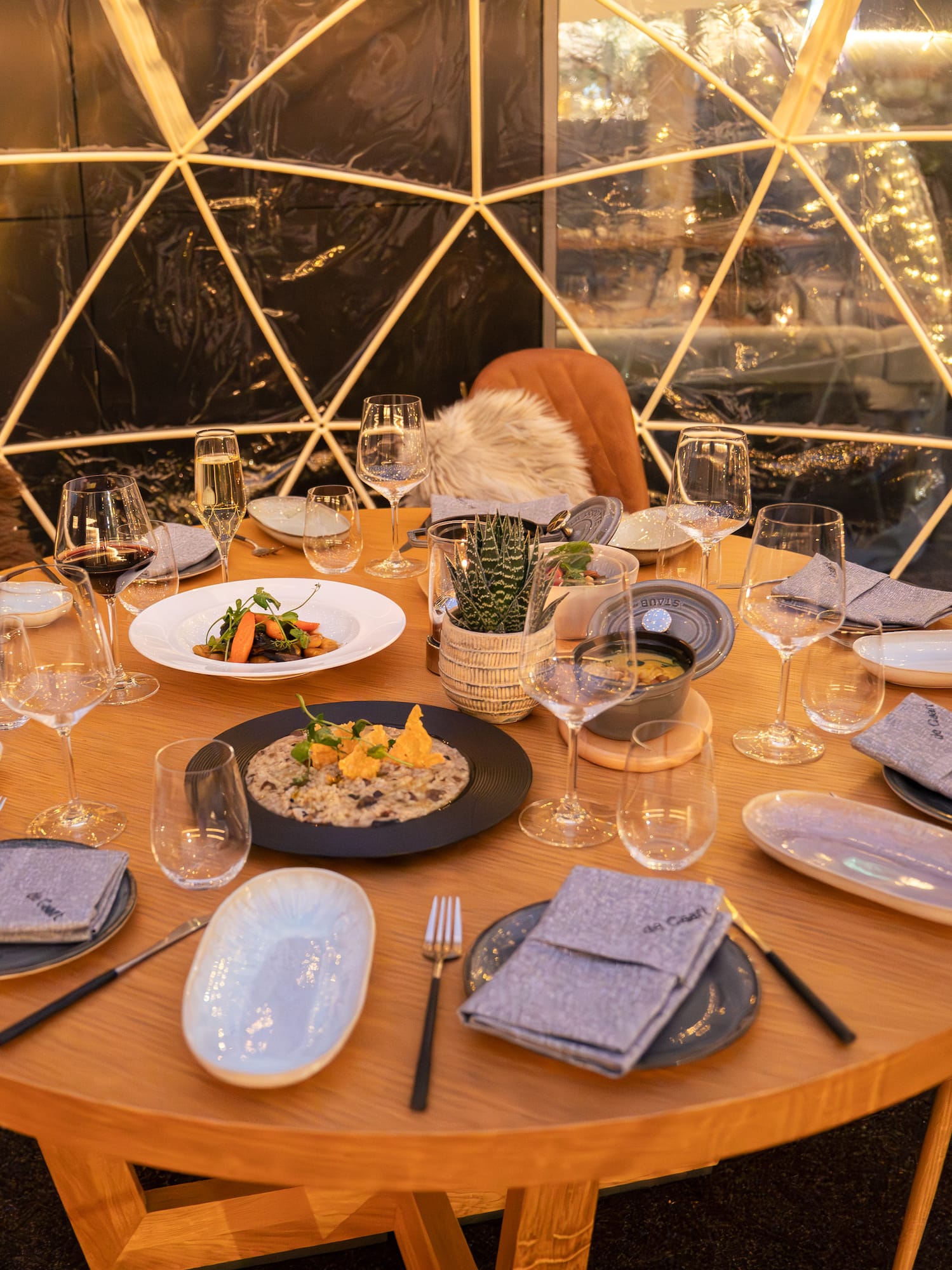 Illuminated dining dome table set with meals, wine, and cozy fur seating.
