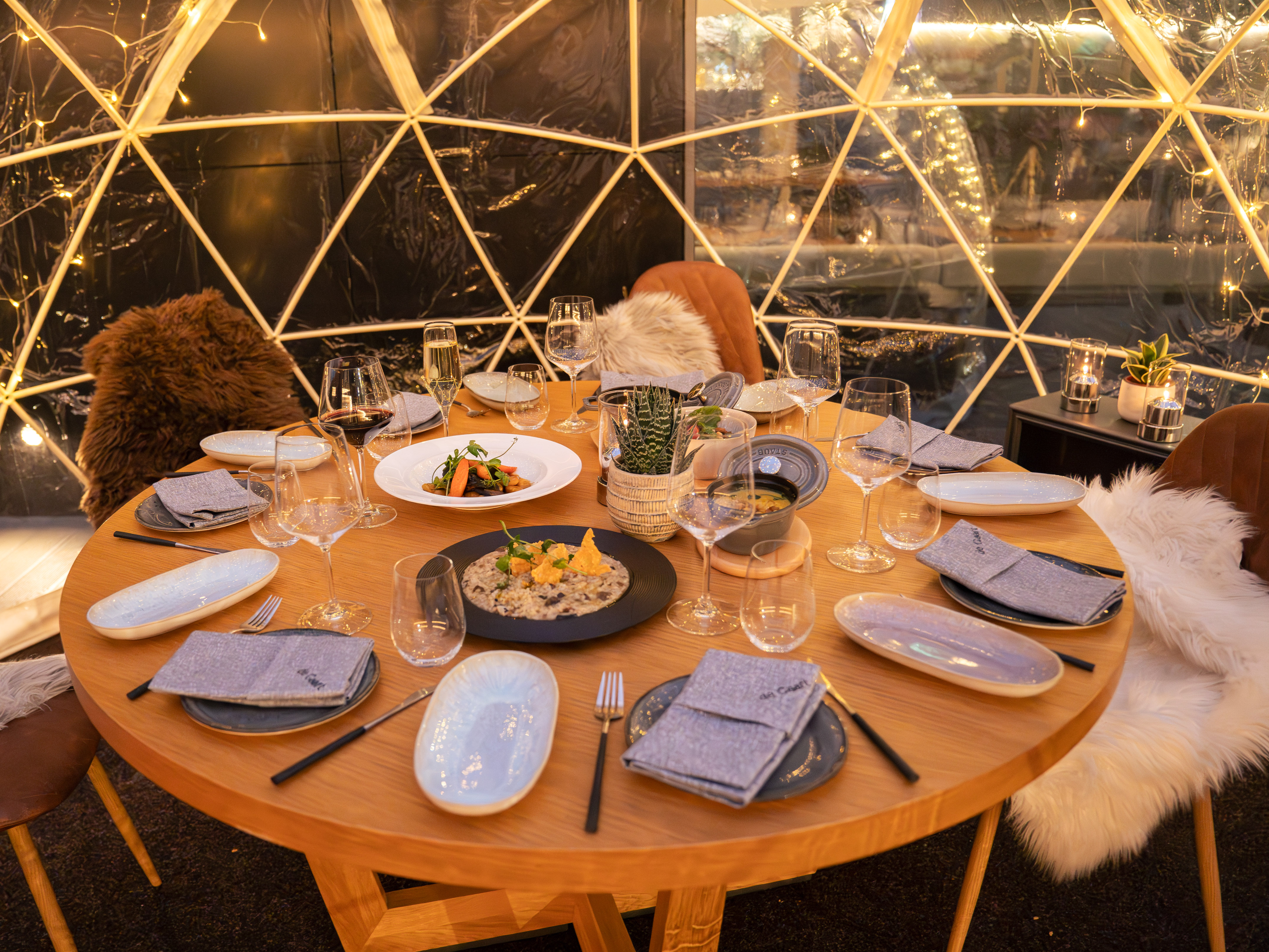 Illuminated dining dome table set with meals, wine, and cozy fur seating.