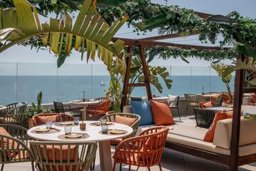 Rooftop restaurant with ocean view, tropical plants, set tables, sailboat.