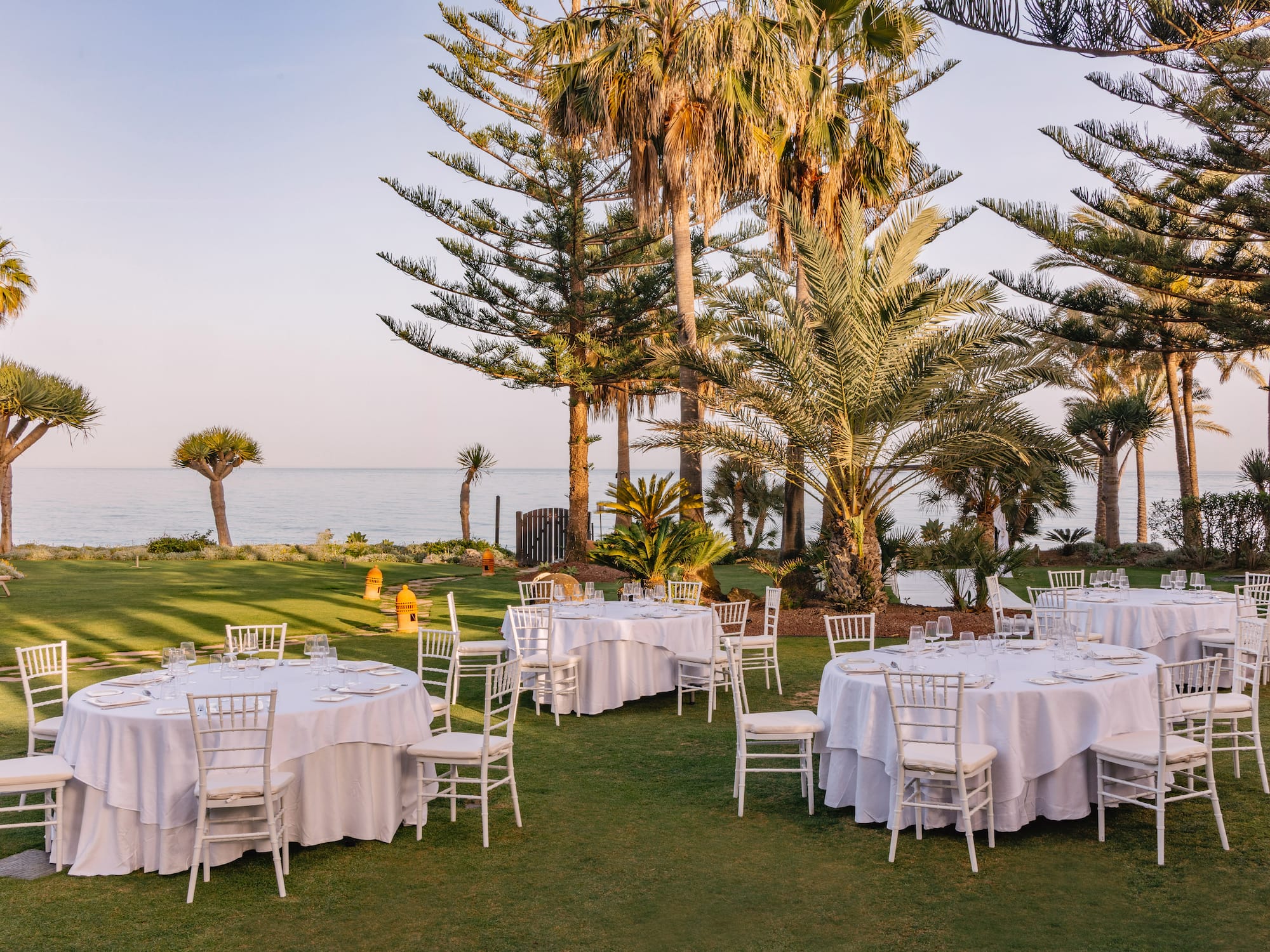 Outdoor dining tables on a green lawn by the ocean, with palm trees.
