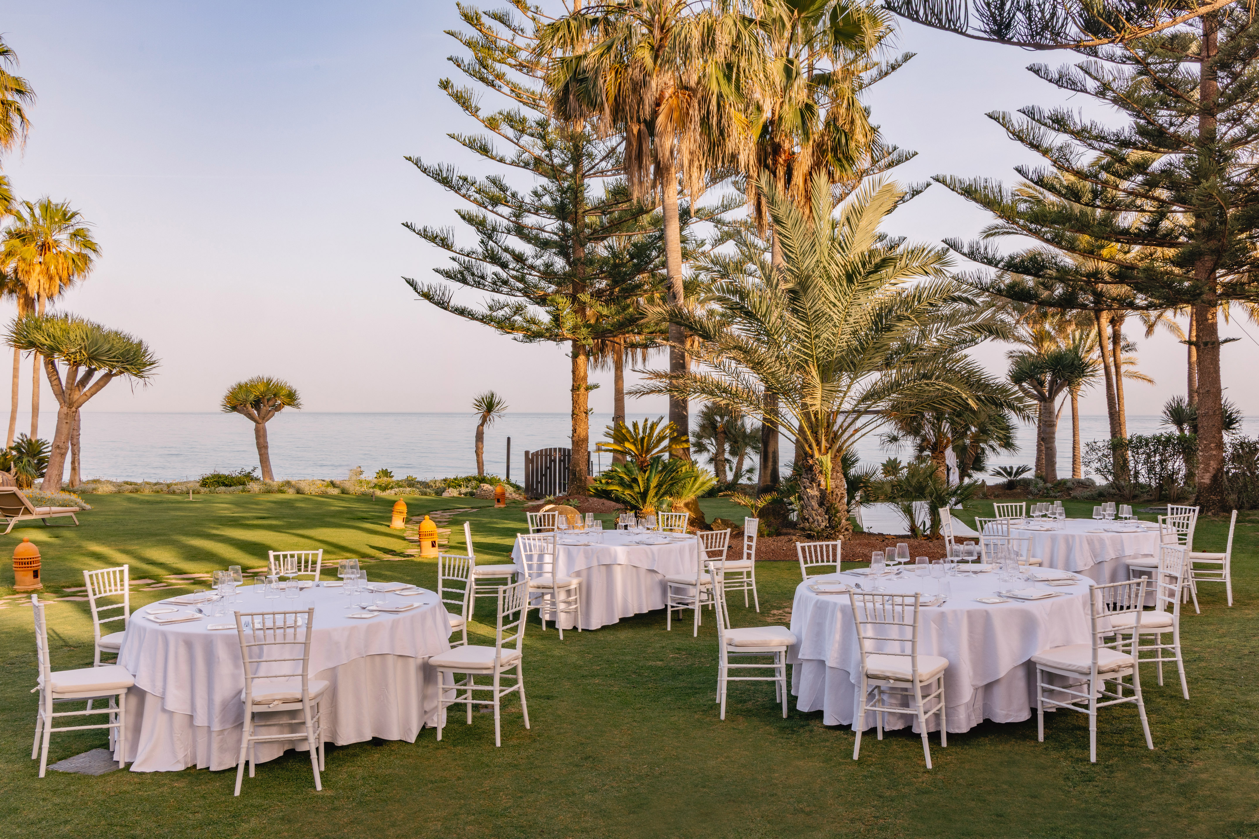 Outdoor dining tables on a green lawn by the ocean, with palm trees.