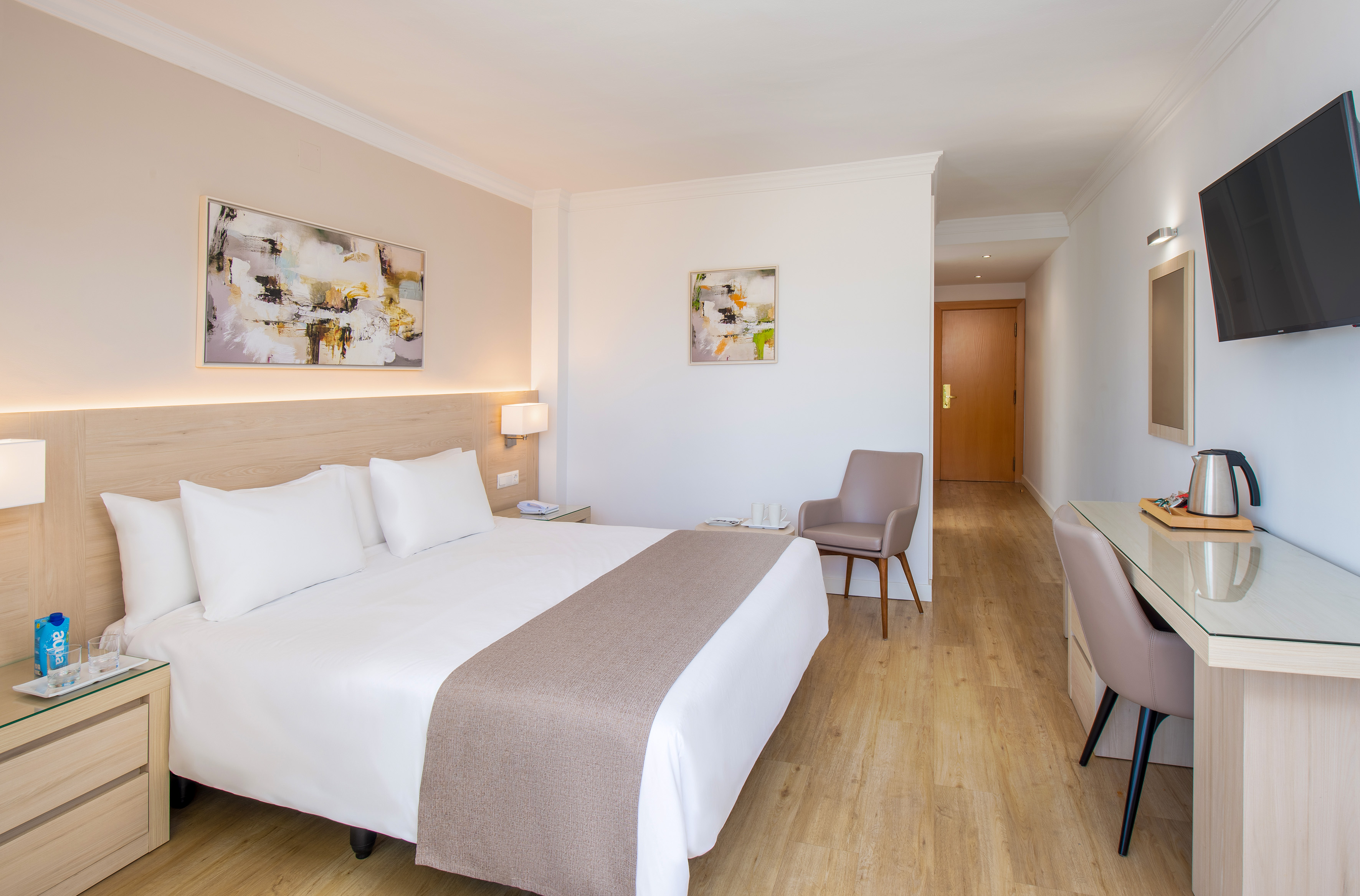Modern hotel guest room with queen bed, desk, TV, and abstract wall art.