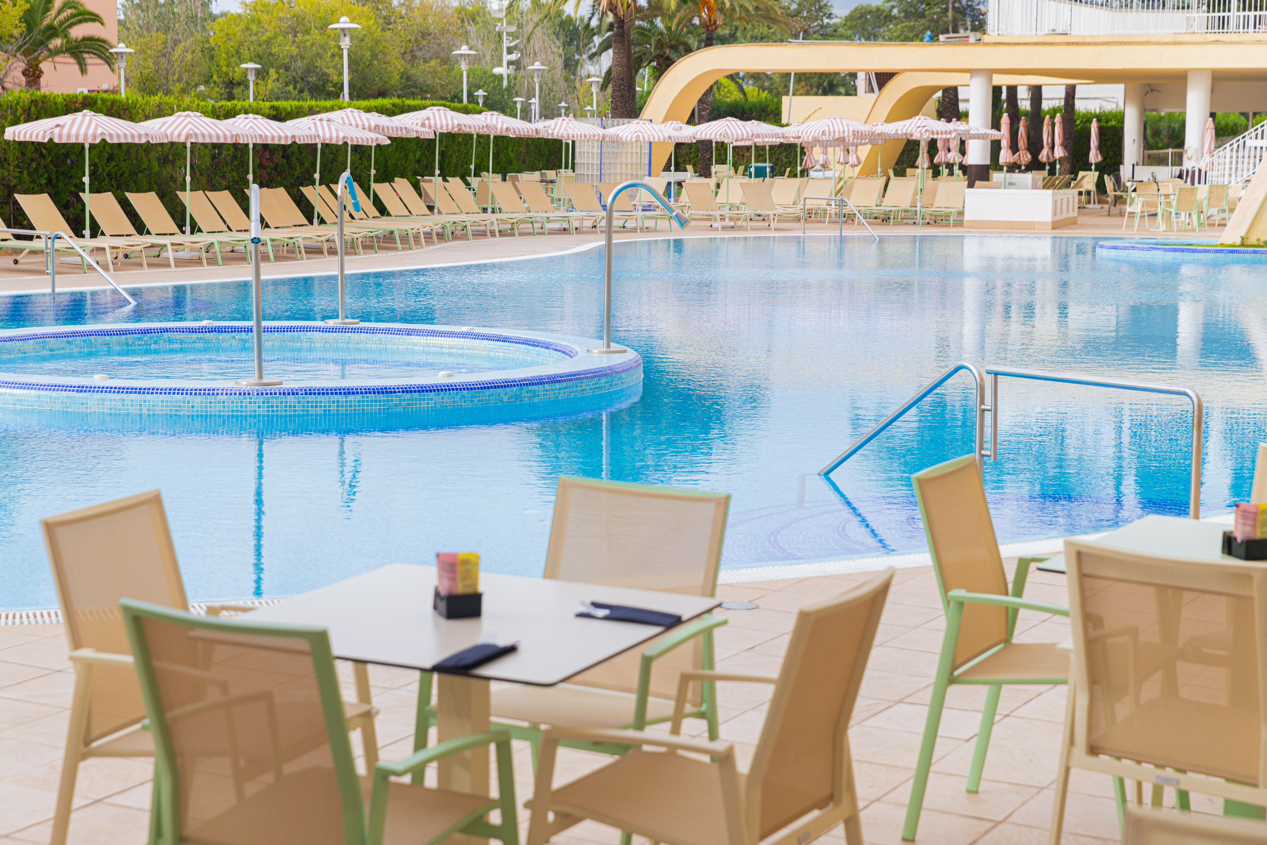 Outdoor hotel pool with sun loungers, striped umbrellas, and poolside cafe tables.