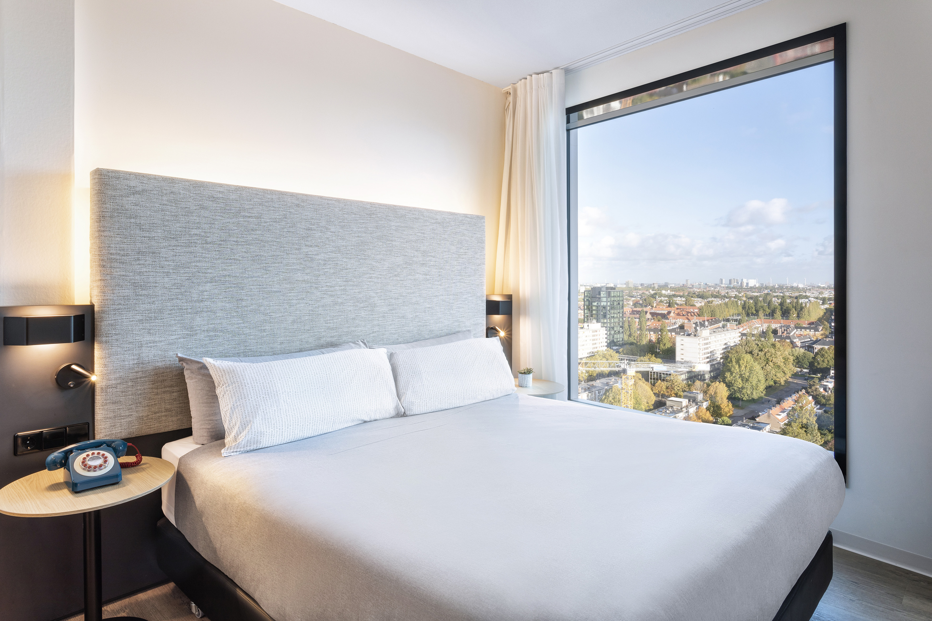 Modern hotel room with large window, city view, grey bed, and vintage blue phone.