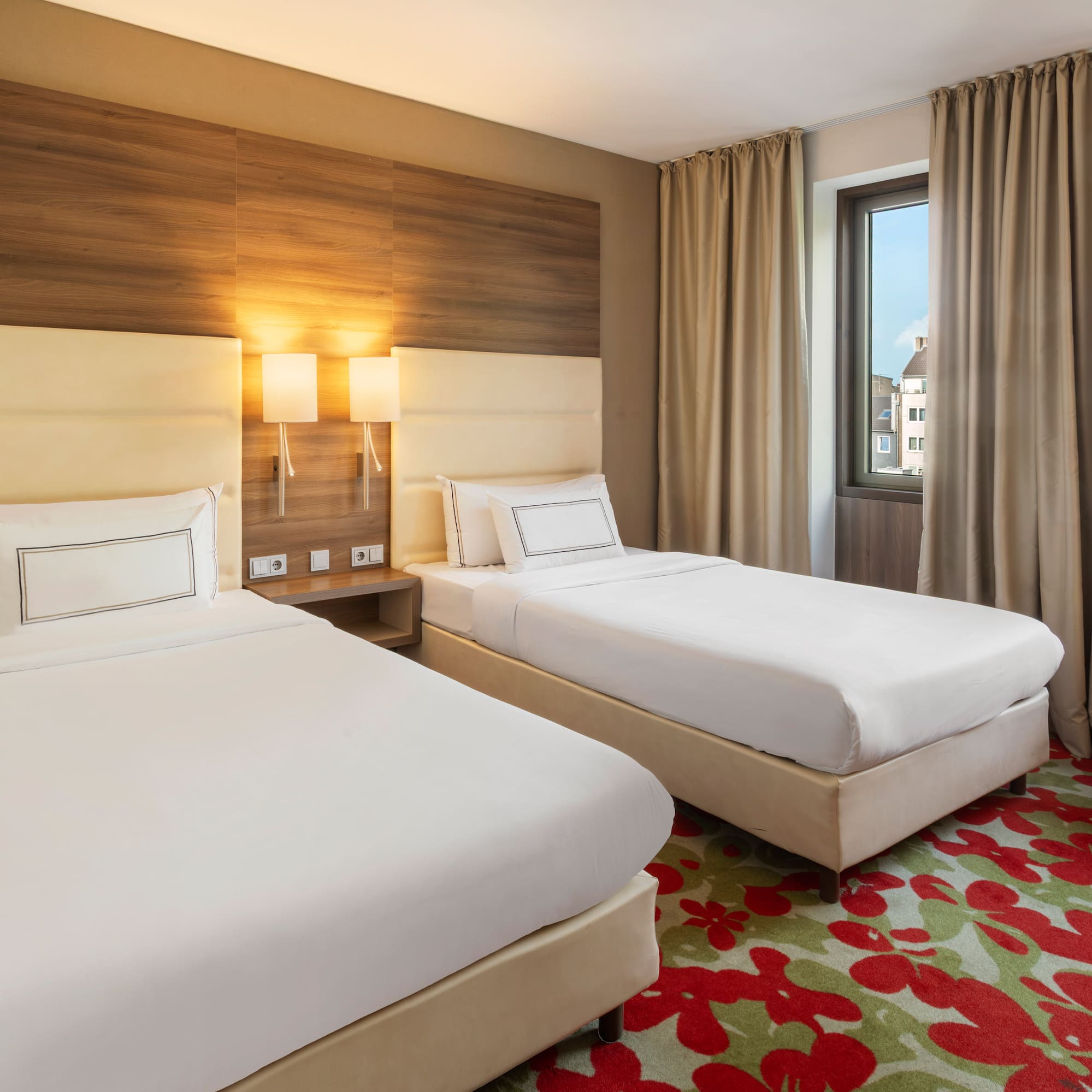 Modern hotel room with two single beds, red armchair, and floral carpet.