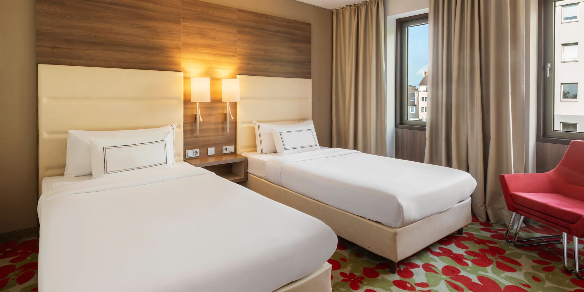 Modern hotel room with two single beds, red armchair, and floral carpet.