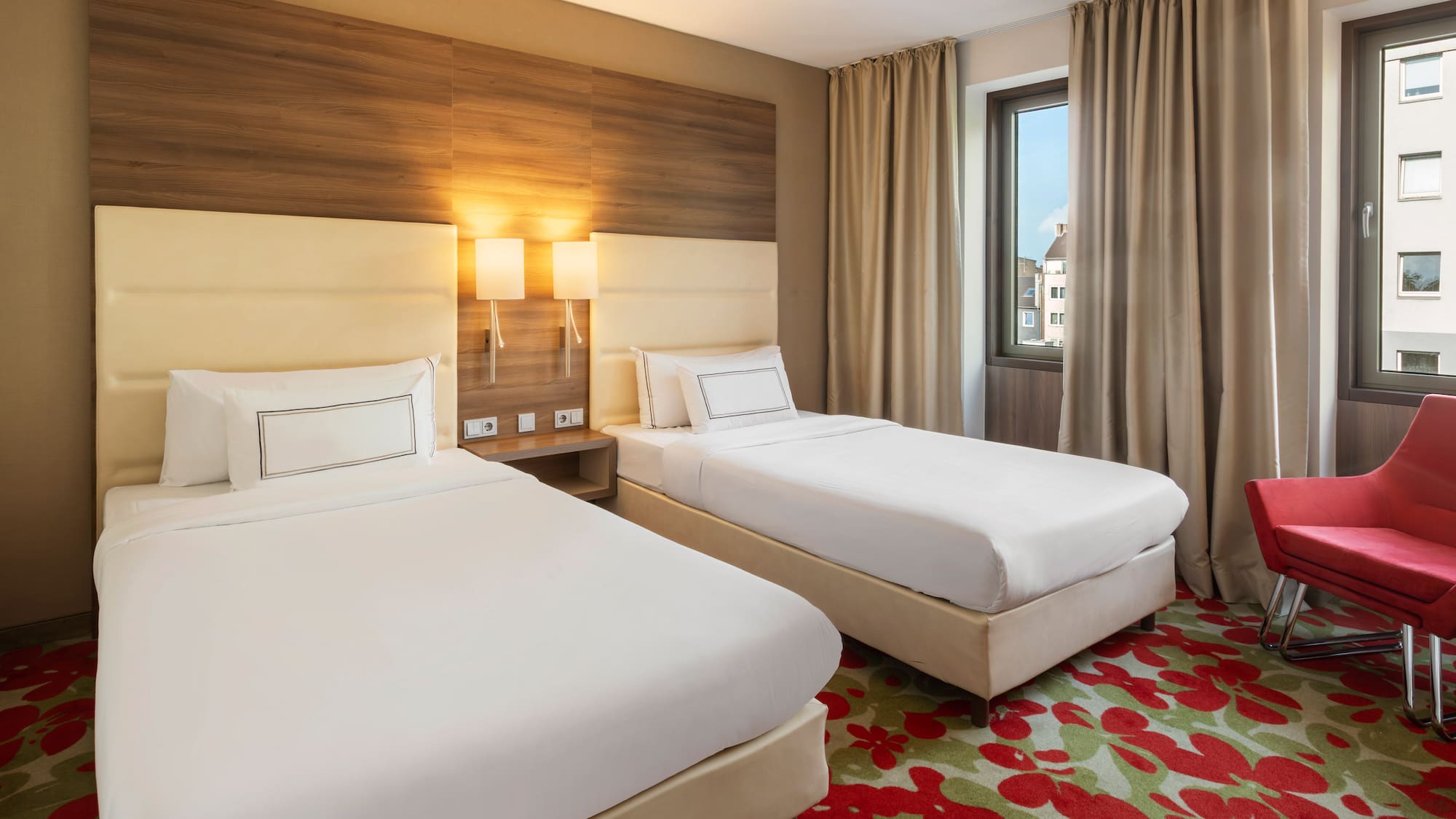 Modern hotel room with two single beds, red armchair, and floral carpet.