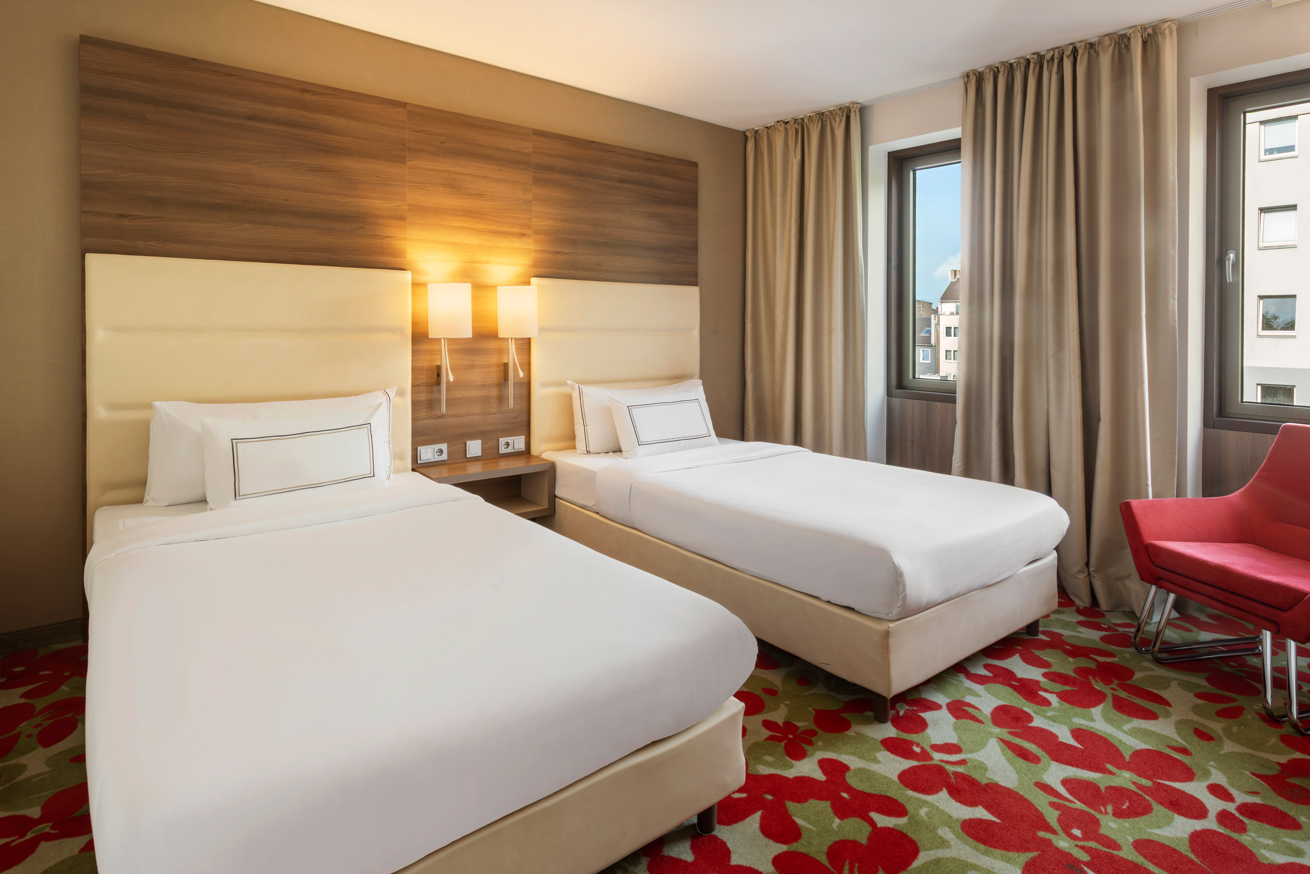 Modern hotel room with two single beds, red armchair, and floral carpet.