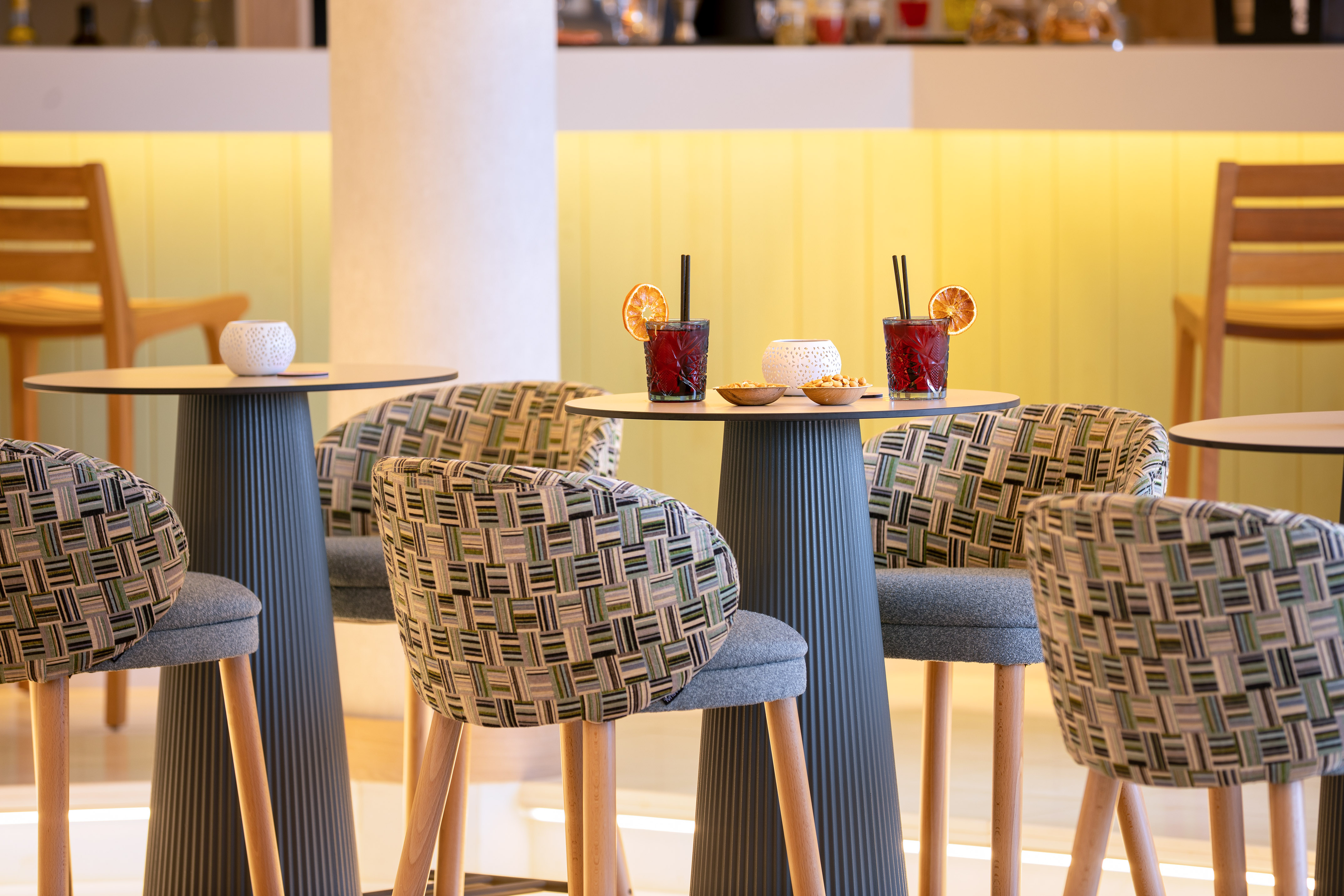 Modern bar interior with red cocktails, patterned high chairs, and yellow lighting.