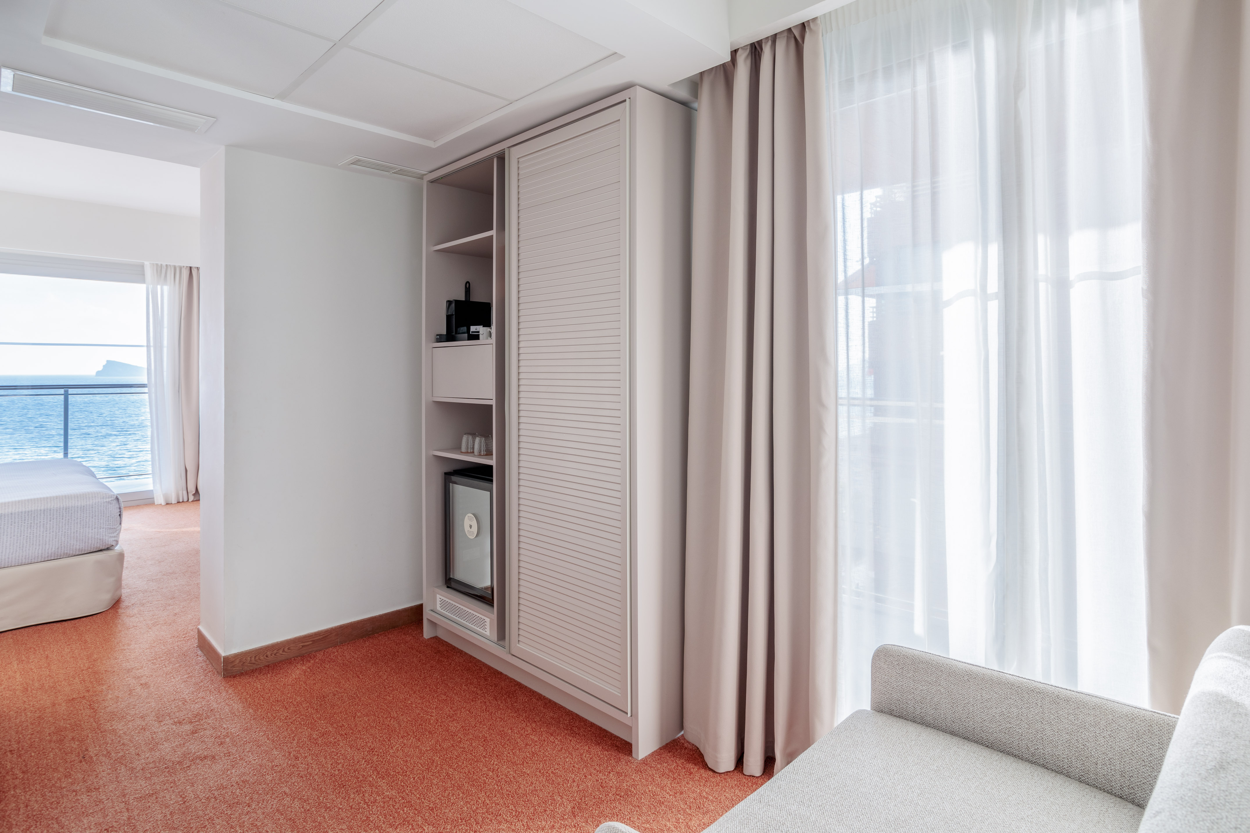 Modern hotel room with sea view, built-in minibar, coffee maker, and orange carpet.