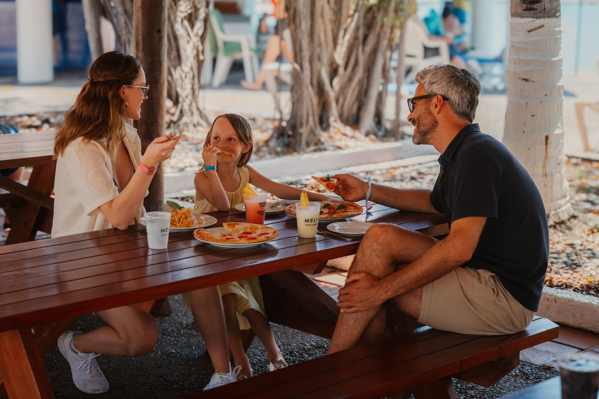 Familia sonriendo comiendo pizza y pasta en resort al aire libre.